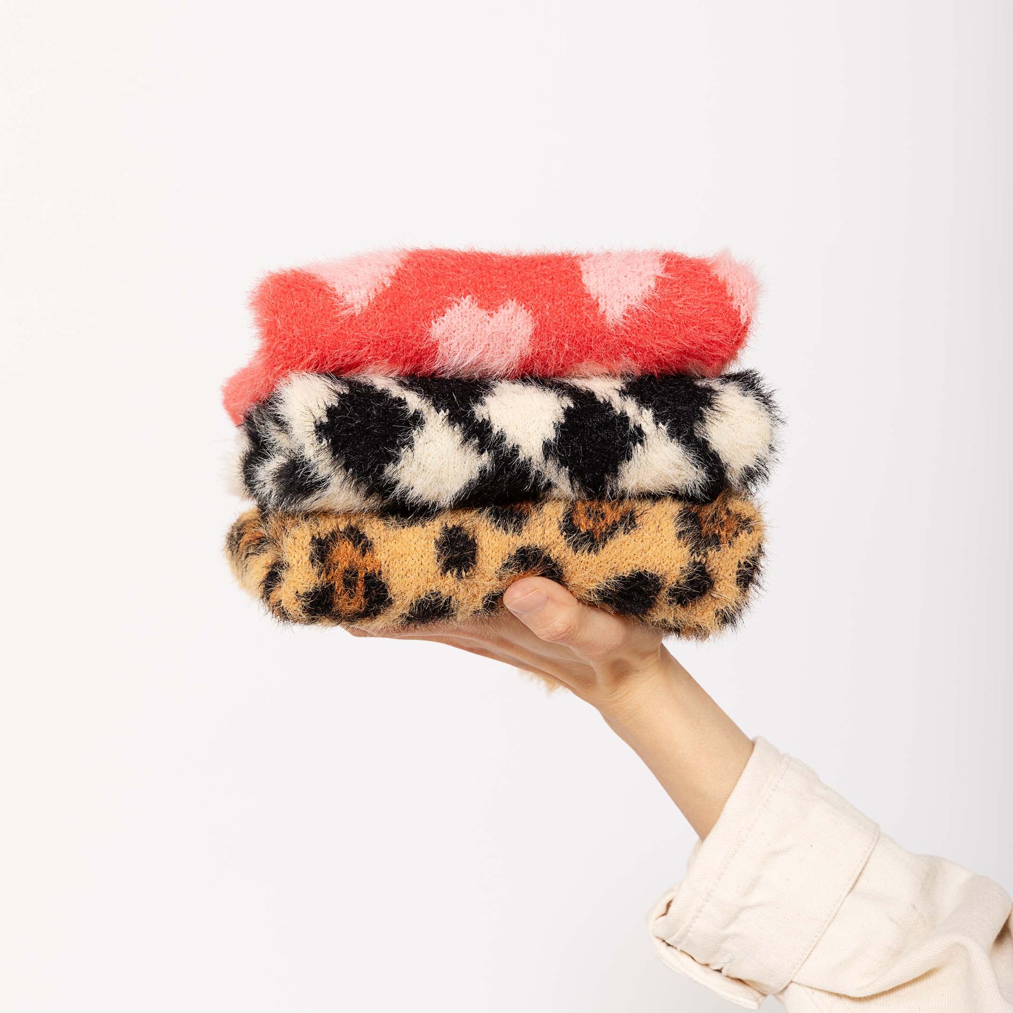 A hand holding a stack of fluffy dog sweaters, with the top one in red with pink hearts and the bottom one in a leopard print, against a white background.