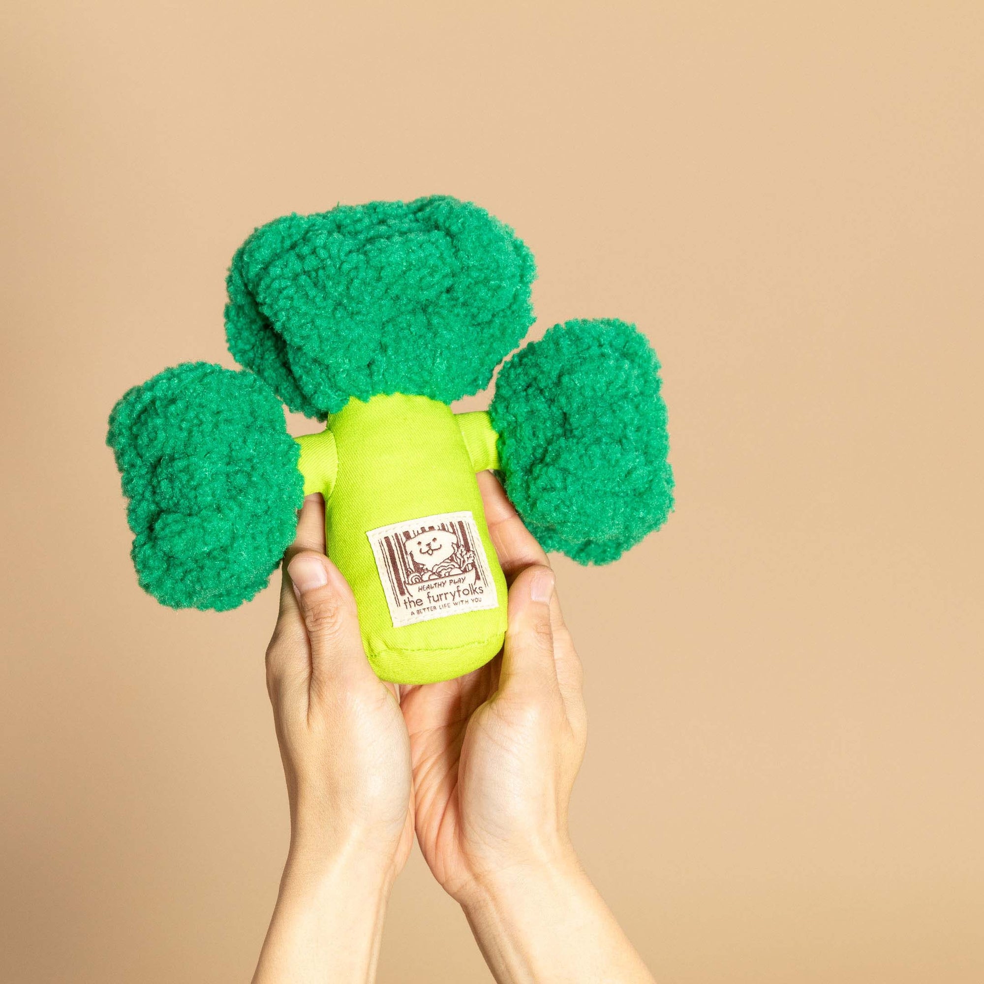Hands holding up a plush broccoli dog toy against a tan background.