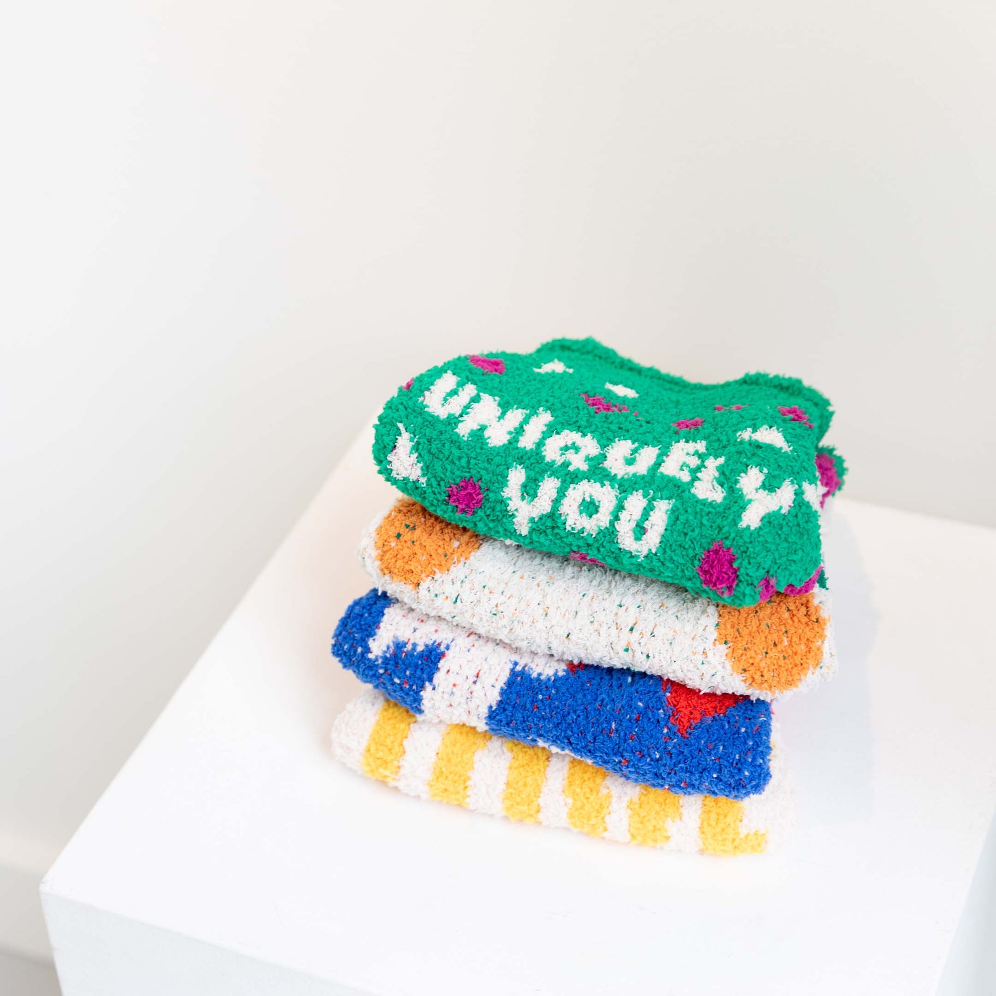 Stack of "The Furryfolks" sweaters with various designs, topped with a green "Uniquely You" sweater on a white cube.