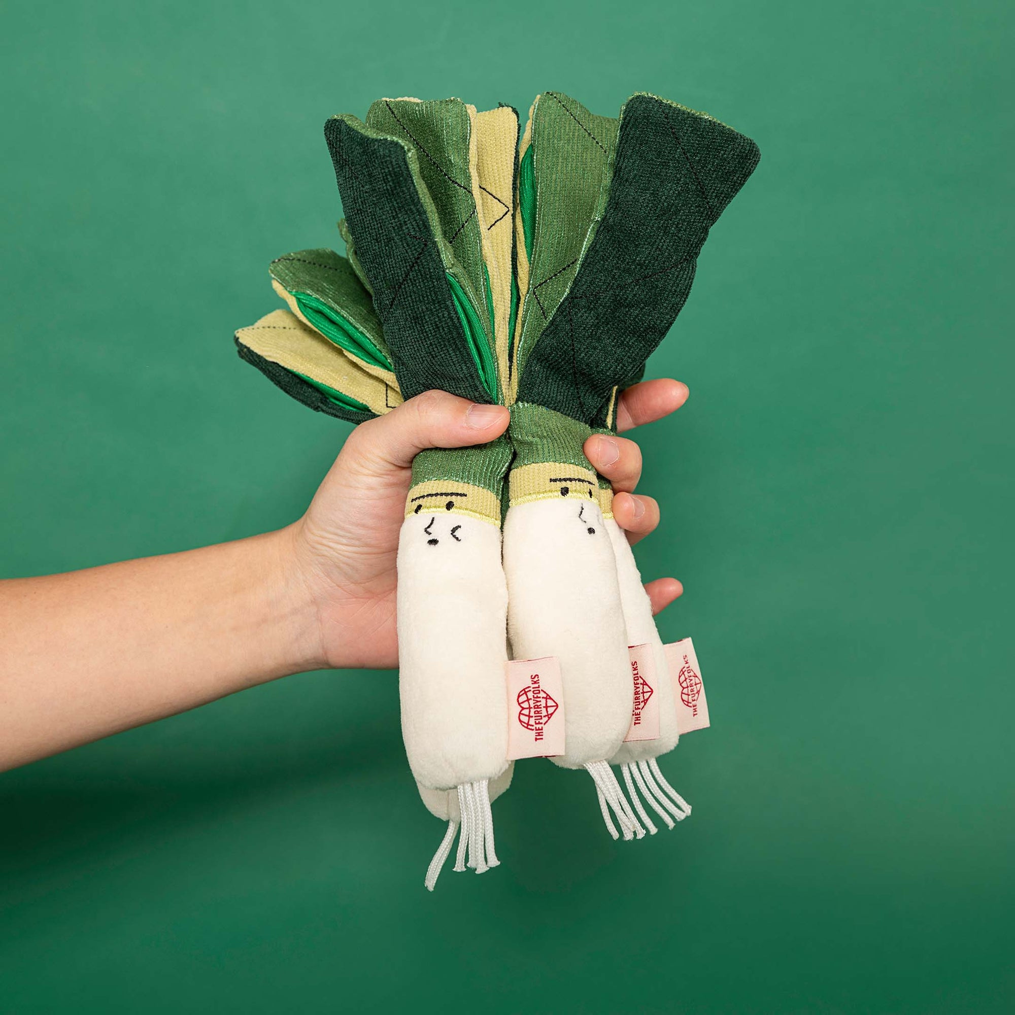 a pair of white and green onion-shaped dog toys being held up against a green background. Each toy has a cute face on the white base and a layered green top representing the onion leaves. The toys are designed for dog engagement, likely intended for interactive play or scent work. The hands holding the toys suggest they are being offered for play, showcasing the interactive nature of the toys.