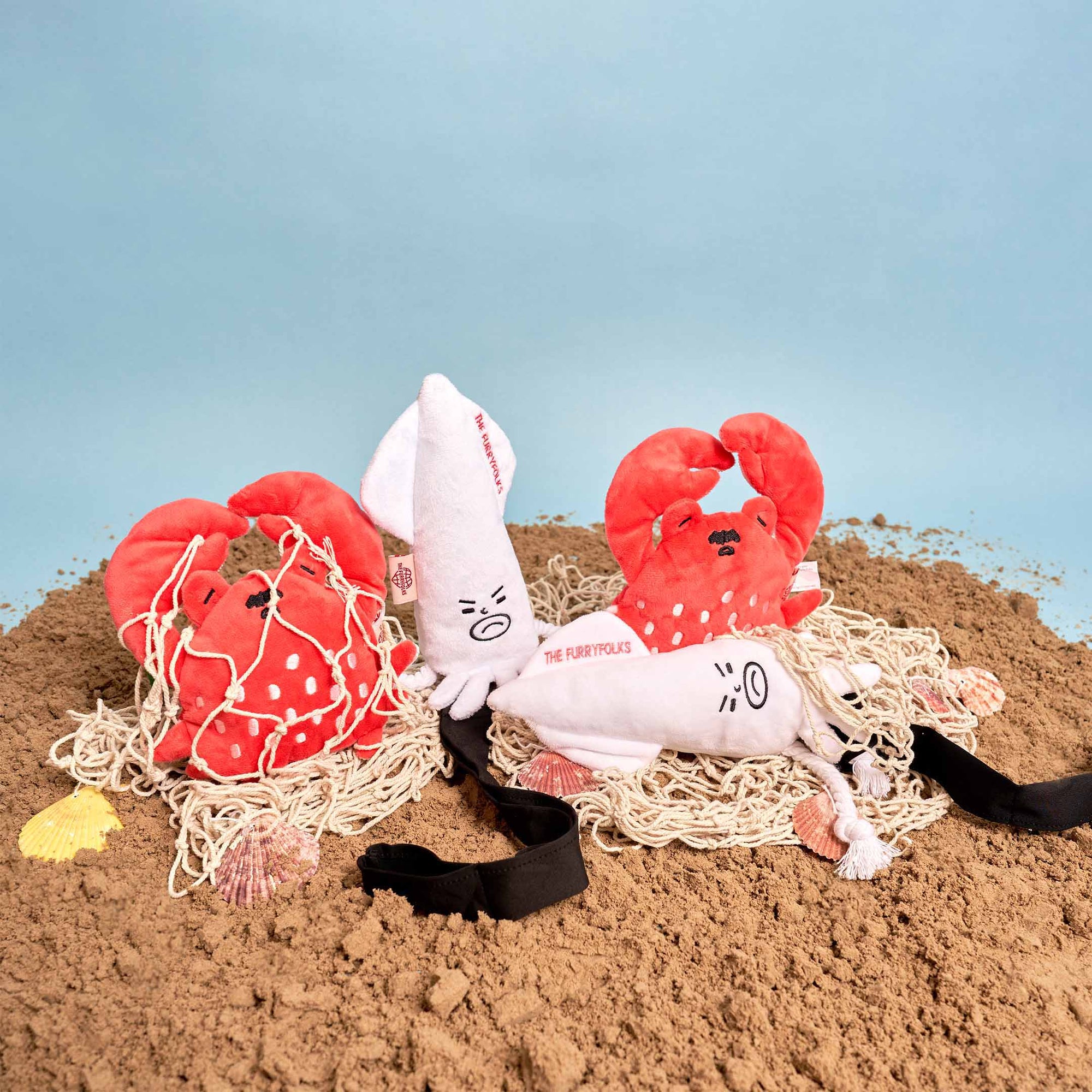 The image showcases a collection of plush toys on a sandy surface, arranged to create a playful, beach-themed setting. The toys include two red crab plushies and a white plush marked with "THE FURRYFOLKS," possibly the brand name. Seashells and a fishing net contribute to the nautical ambiance. This arrangement could be for product promotion or a creative display concept.