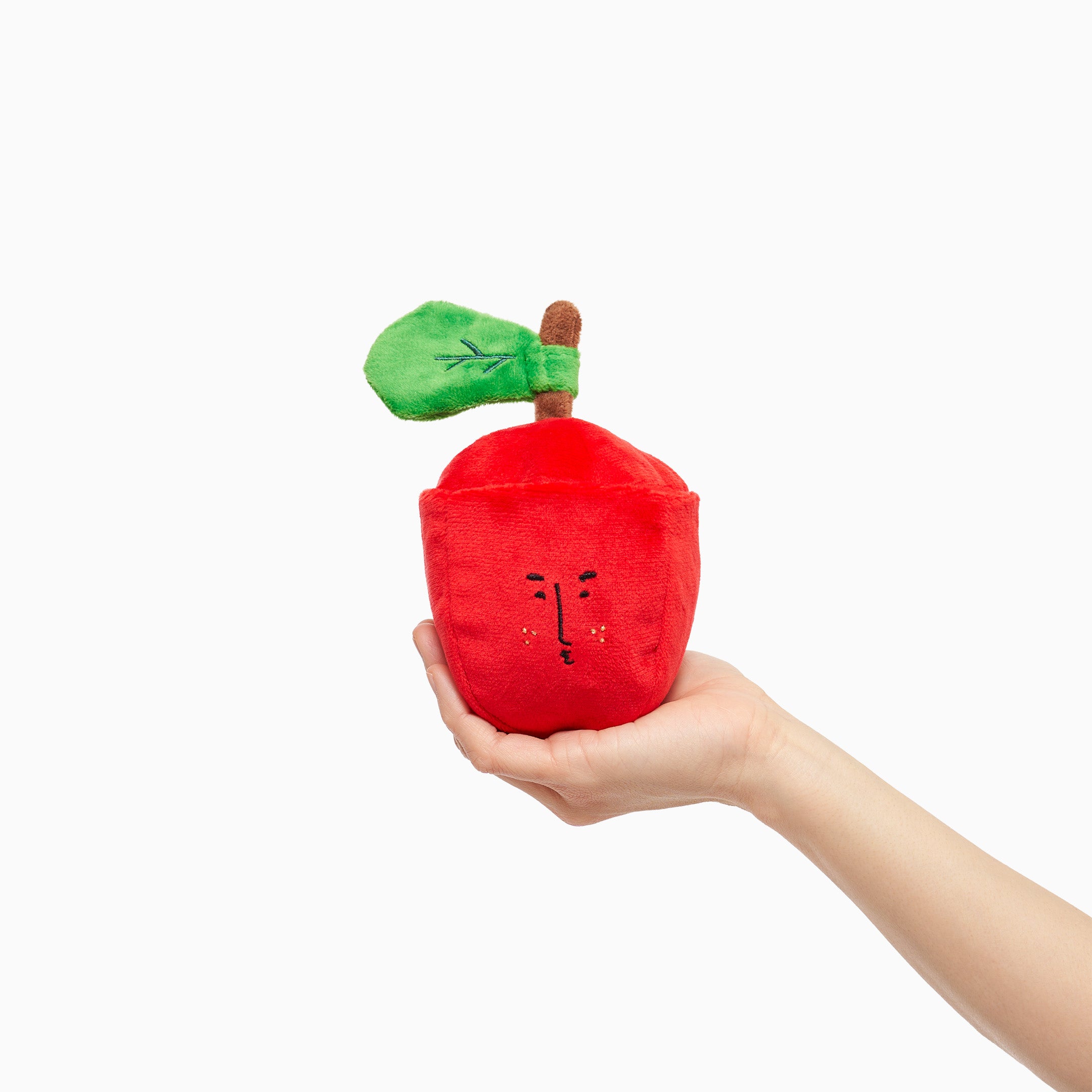 A person's hand holding a red apple-shaped dog toy with a cute face and a green leaf on white background.
