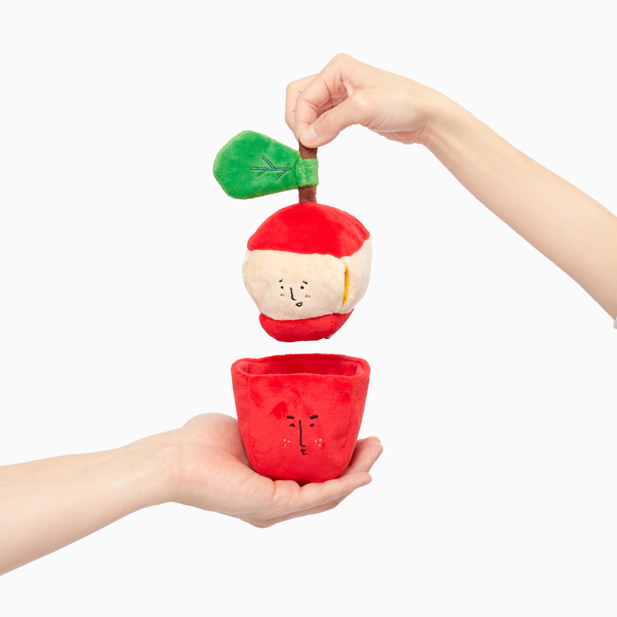 Two hands holding parts of a red and white apple-shaped dog toy with a cute face; one hand holds the top with the green leaf, and the other hand holds the bottom on a white background.