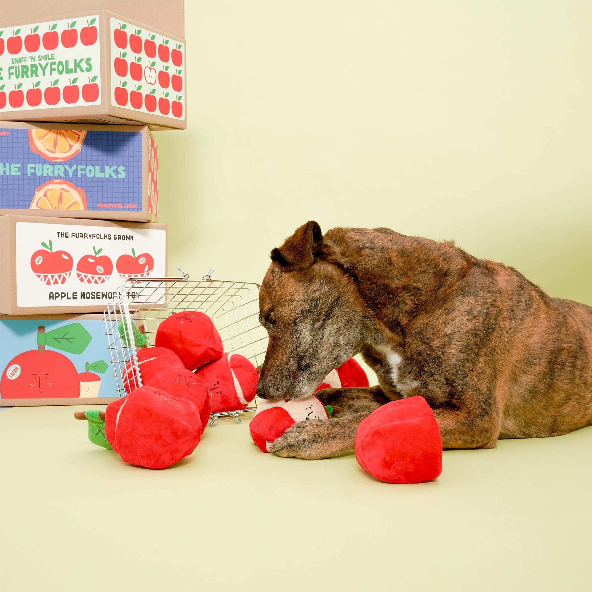 A brown dog examining red apple-shaped dog toys spilled from a tipped-over shopping cart, with boxes labeled "The Furryfolks Apple Nosework Toy" in the background.