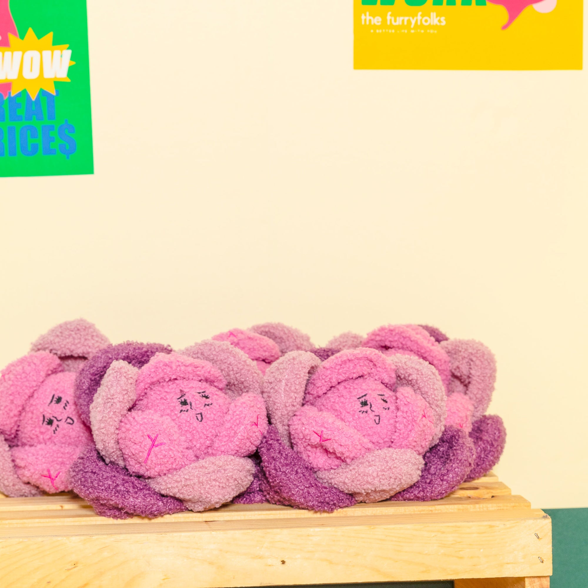 The photo shows a collection of plush Red Cabbage toys in pink and purple, arranged on a wooden platform with promotional posters in the background. The posters feature phrases like "WOW GREAT PRICES" and branding for "the furryfolks", hinting at a sale or event for pet products. The setting suggests a fun and engaging shopping experience aimed at pet owners. The beige and green wall colors complement the toys' vibrant hues, creating a cheerful and inviting atmosphere.