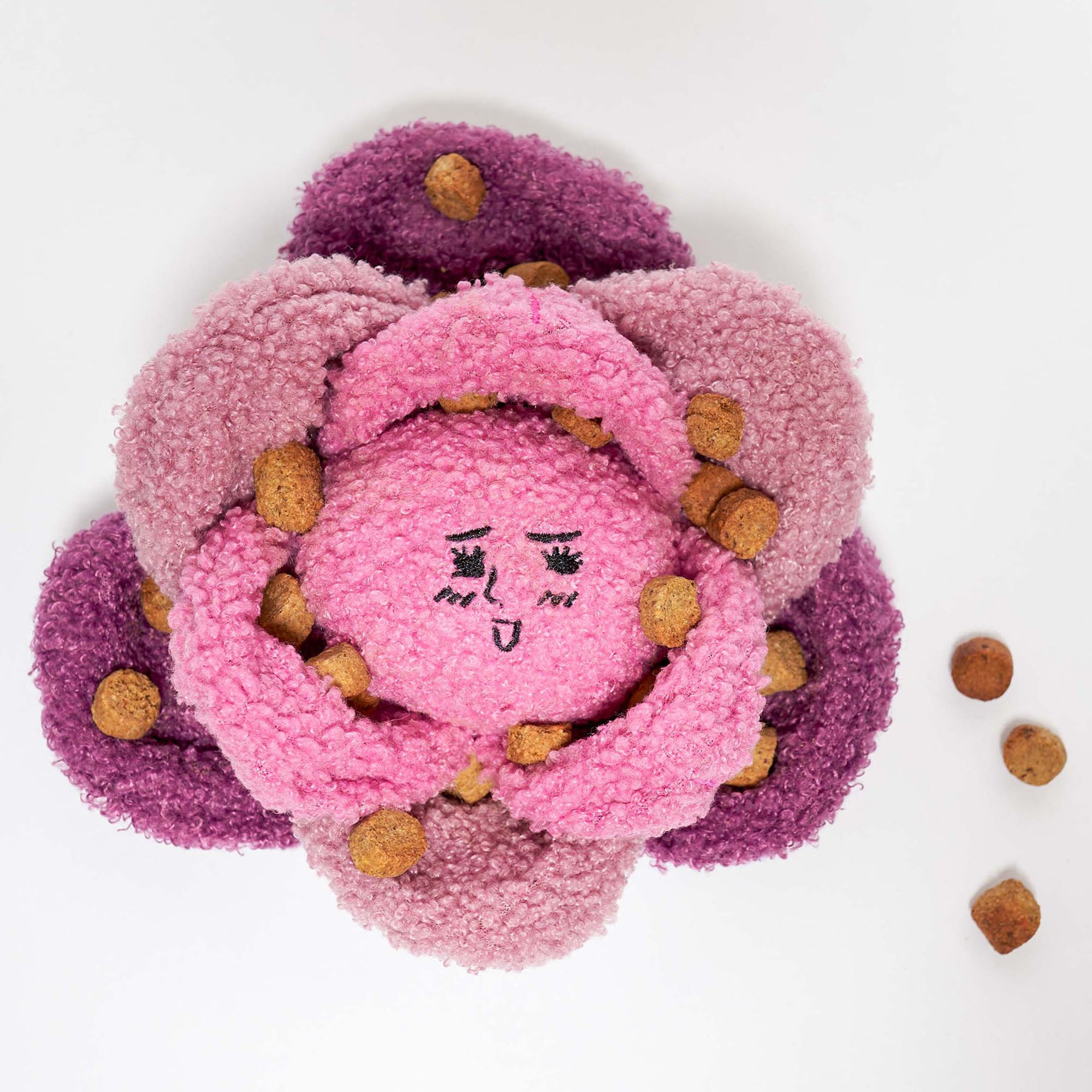 The image shows a dog nosework toy resembling a red cabbage, with multiple fabric layers in pink and purple, and brown elements mimicking seeds. The smiling face at the center adds charm. This interactive toy stimulates a dog's sense of smell, with pockets for hiding treats. The plain background highlights the toy's design.