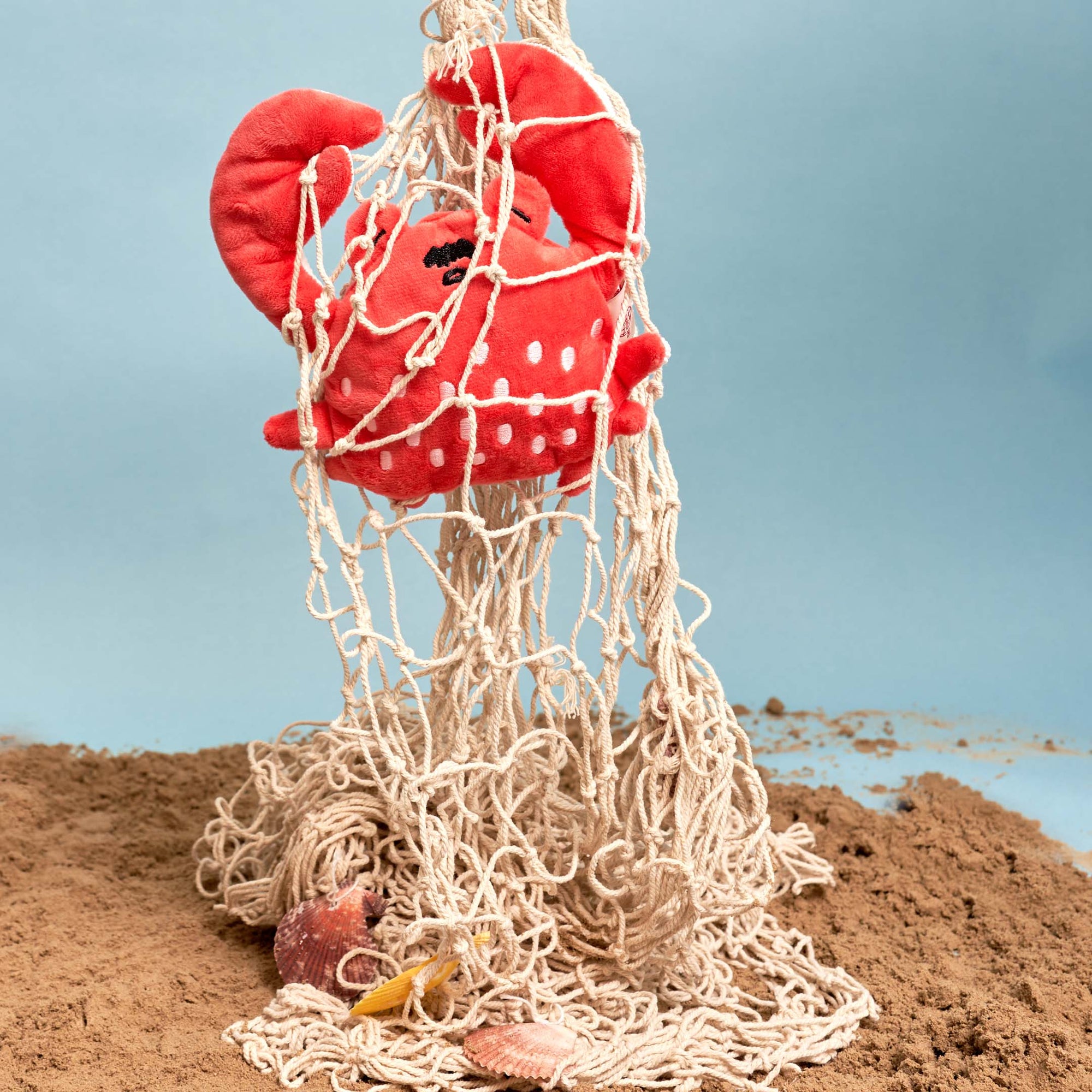 The image shows the plush red crab toy entangled in a fishing net, with a backdrop suggesting a sandy, beach-like environment. There are also shells and starfish partially buried in the sand at the base of the net, adding to the maritime theme. This setting is possibly staged to evoke the natural habitat of a crab, creating a playful and thematic presentation of the toy, which might be used for promotional purposes or as a creative display.