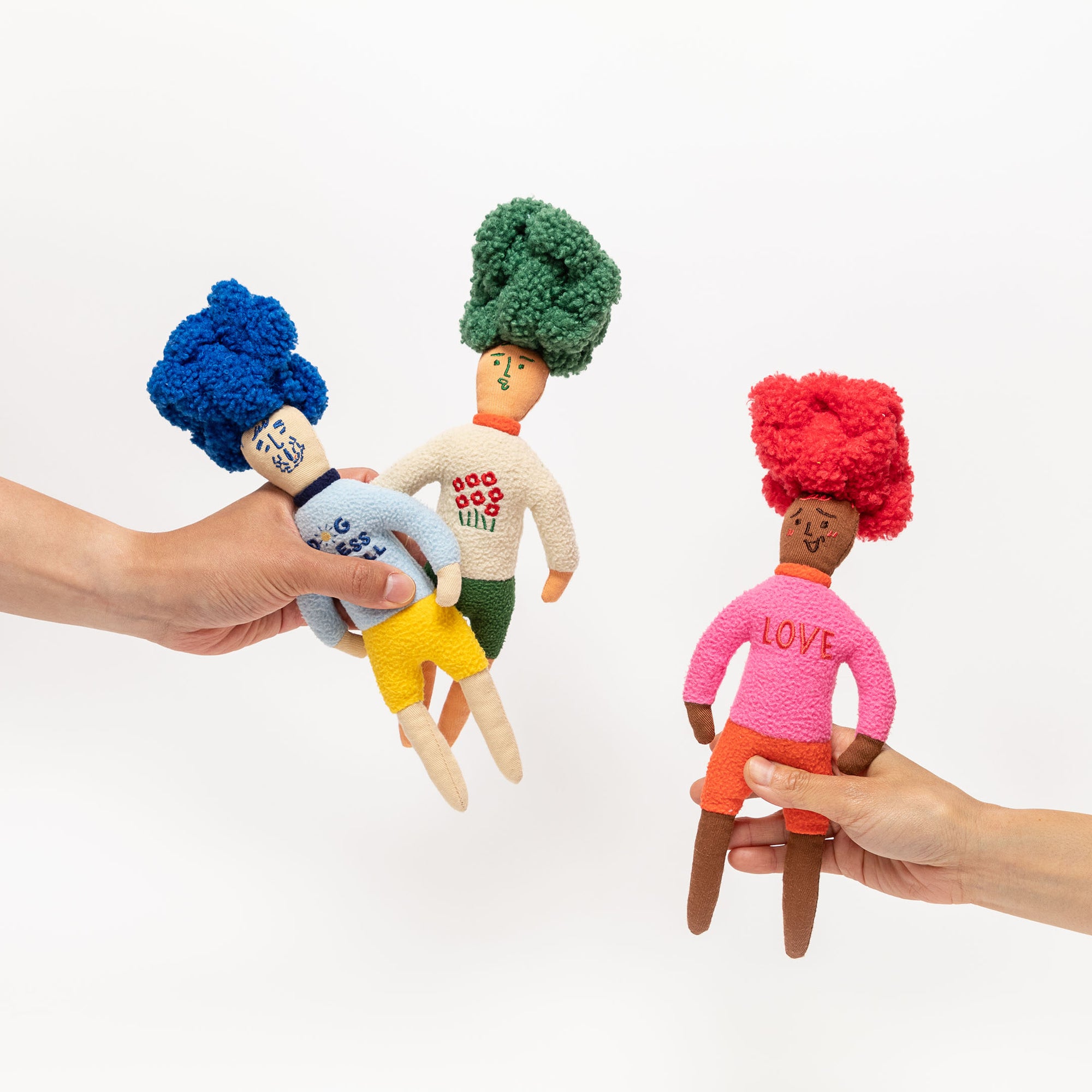  The image shows three nosework dog toys designed as colorful, handcrafted dolls with yarn hair in blue, green, and red. They are made with textured fabrics and have features that might hide treats to stimulate a dog's sense of smell. Each toy has a distinct look, with one wearing a cloud-patterned shirt, another with polka dots, and the third with "LOVE" on its shirt. They serve both as engaging sensory toys for pets and as playful decor items.