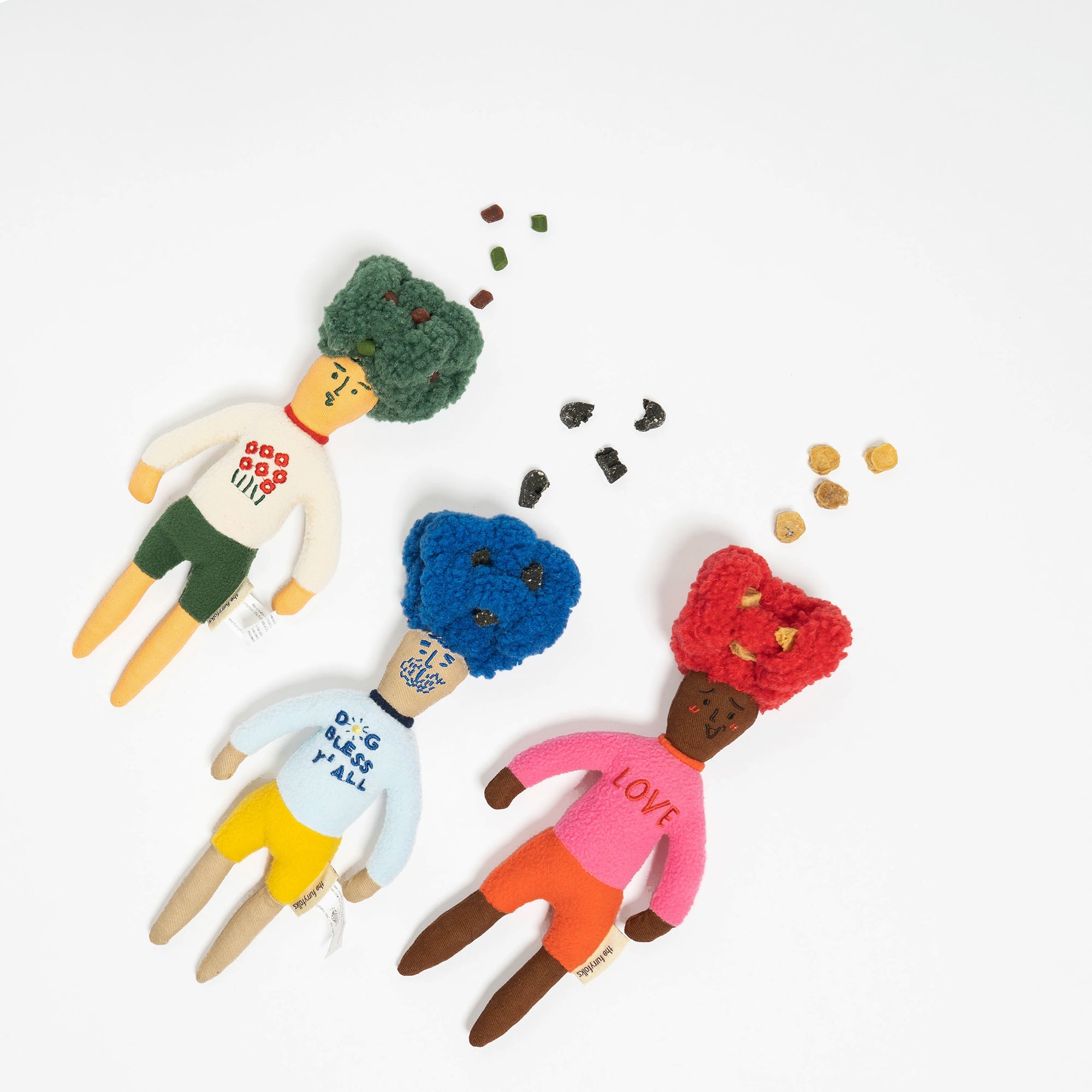 The image shows three dog toys designed as dolls with colorful yarn hair, each paired with different treats on a white background. These toys are for nosework training, allowing dogs to sniff out the treats hidden inside. With green, blue, and red hair, and treats scattered around, they combine functionality with a playful aesthetic, perfect for engaging a dog's sense of smell and providing mental stimulation.