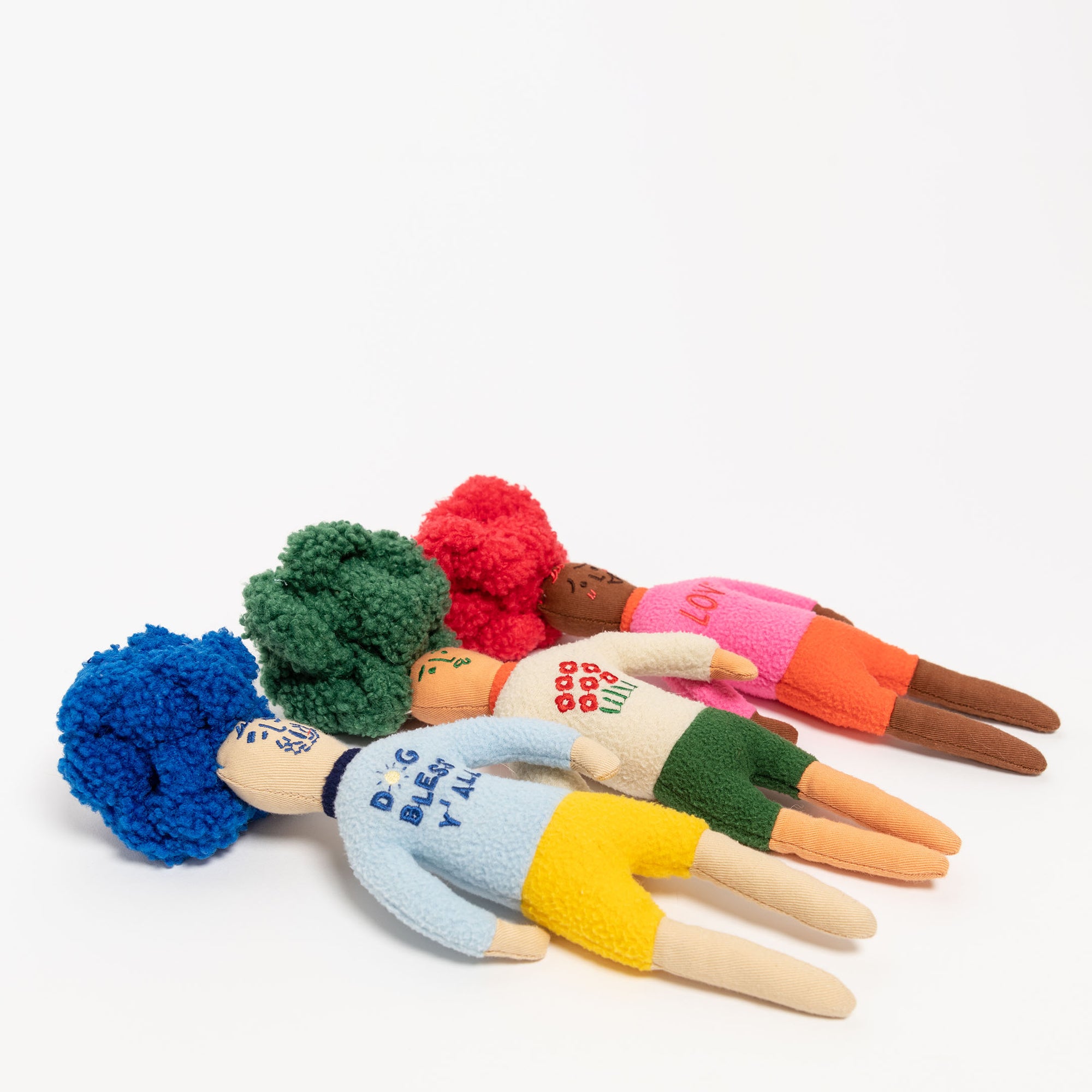 The image displays a set of four handcrafted nosework toys, designed as colorful dolls with pom-pom hair in shades of blue, green, red, and brown, set against a white background. Each toy has unique clothing—a blue sweater with "DOG BLESS Y'ALL," a green one with red polka dots, a pink "LOVE" shirt, and a cream with red dots—meant to hide treats for dogs to find. These toys combine functionality and style, catering to a dog's sense of smell and the owner's aesthetic taste.