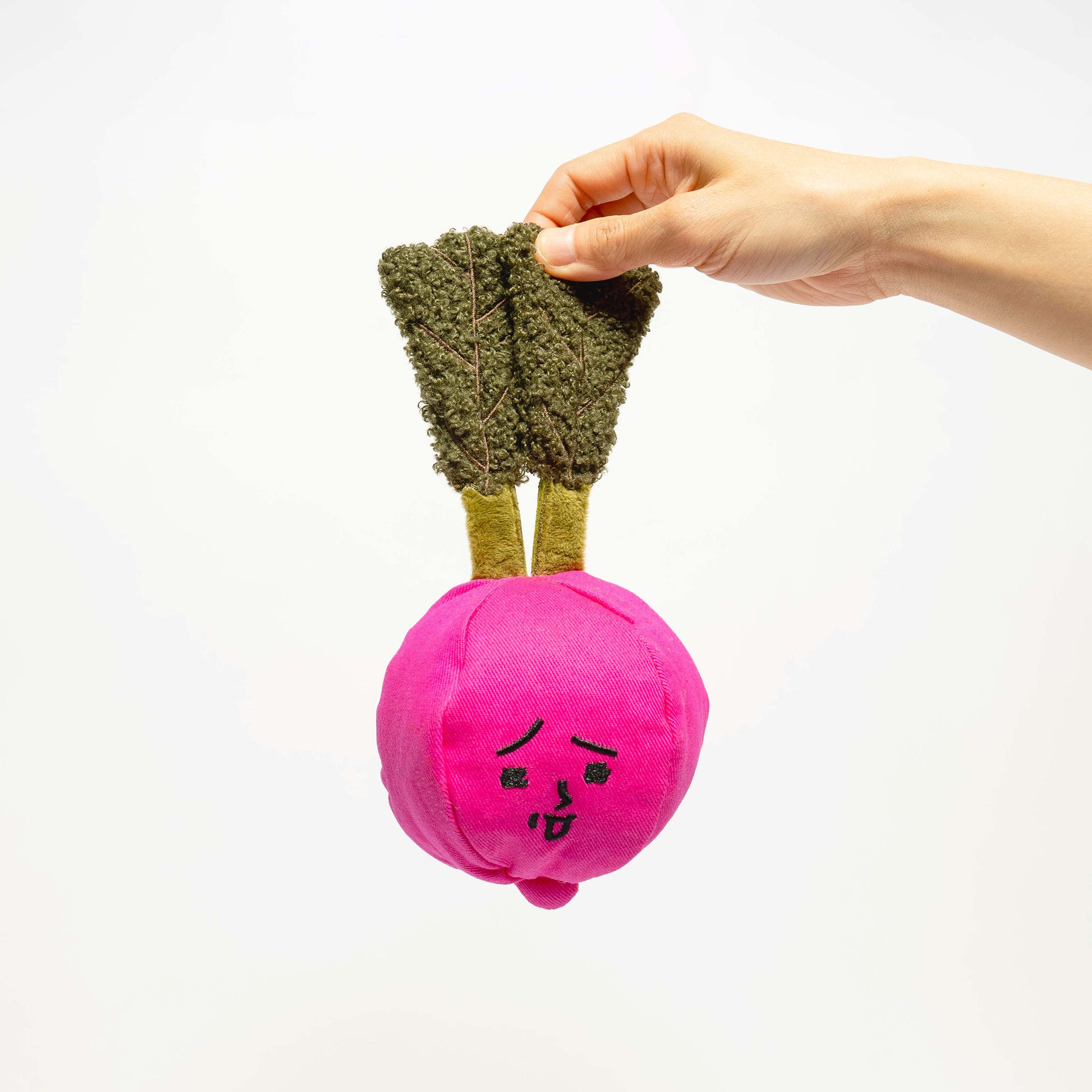 The image features a hand holding a unique dog toy shaped like a radish. The toy has a bright pink bulb with a cute, sleepy face embroidered on it and green fabric leaves that mimic the appearance of a real radish top. The texture of the leaves looks soft and is likely designed for a dog's tactile enjoyment, while the overall shape and color of the toy make it visually appealing and easy for both dogs and owners to find during play. The white background enhances the vivid colors and detail of the toy.