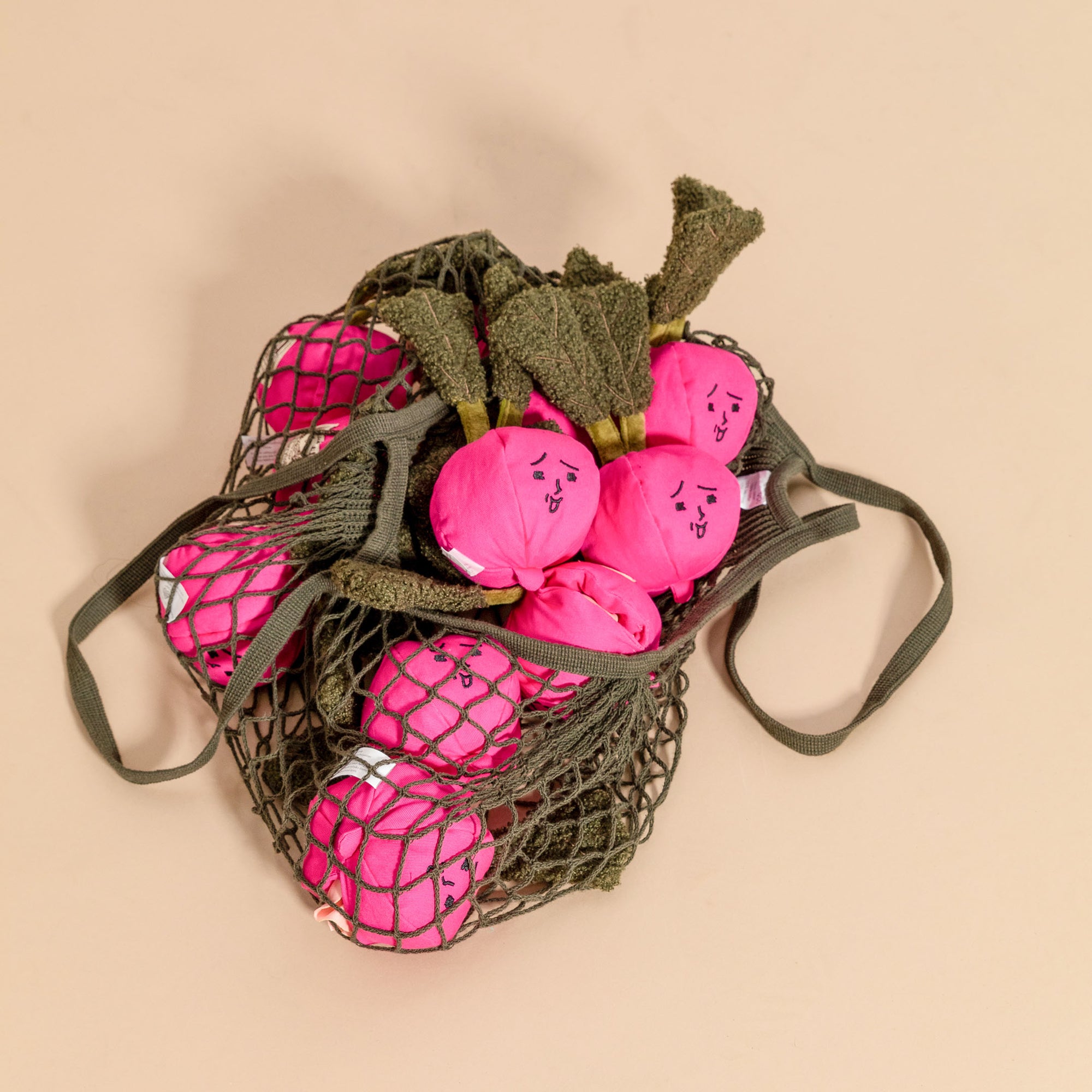 The image displays a collection of pink radish-shaped dog toys with sleepy faces, nestled in a green net bag. The toys are designed with bright pink bulbs and green tops to closely resemble real radishes. This playful design is both visually charming and likely to be engaging for pets, with the net bag adding a practical element for storage and transport. The toys are set against a peach-colored background, which complements the toys' colors and creates a warm, inviting aesthetic.