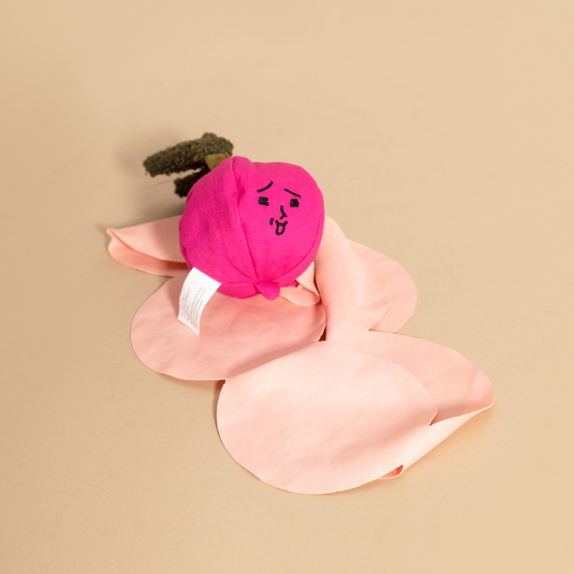The image shows a pink radish-shaped dog toy with a sleepy face and leaf-like extensions, set against a tan background. It features a vibrant pink bulb and contrasting green top, resembling a radish sprouting from the ground. The light pink elements suggest petals, adding a whimsical touch to the toy's design. A visible tag indicates the toy's commercial branding or care instructions, emphasizing its crafted quality.