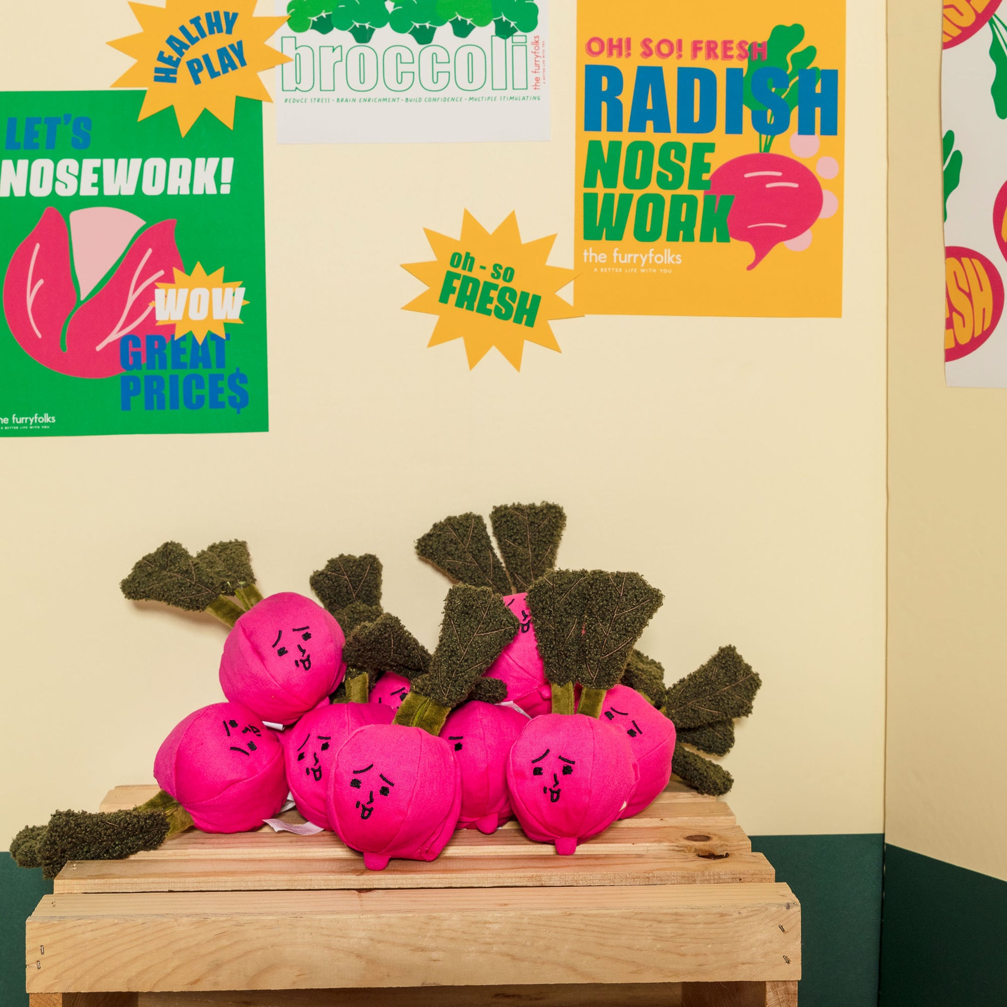 The image shows a playful setup with pink radish-shaped dog toys on a wooden stand against a backdrop of colorful posters. The posters advertise nosework toys with catchy phrases like "Healthy Play" and "Oh So Fresh". This scene is likely part of a pet store or event promoting interactive and enriching activities for dogs.