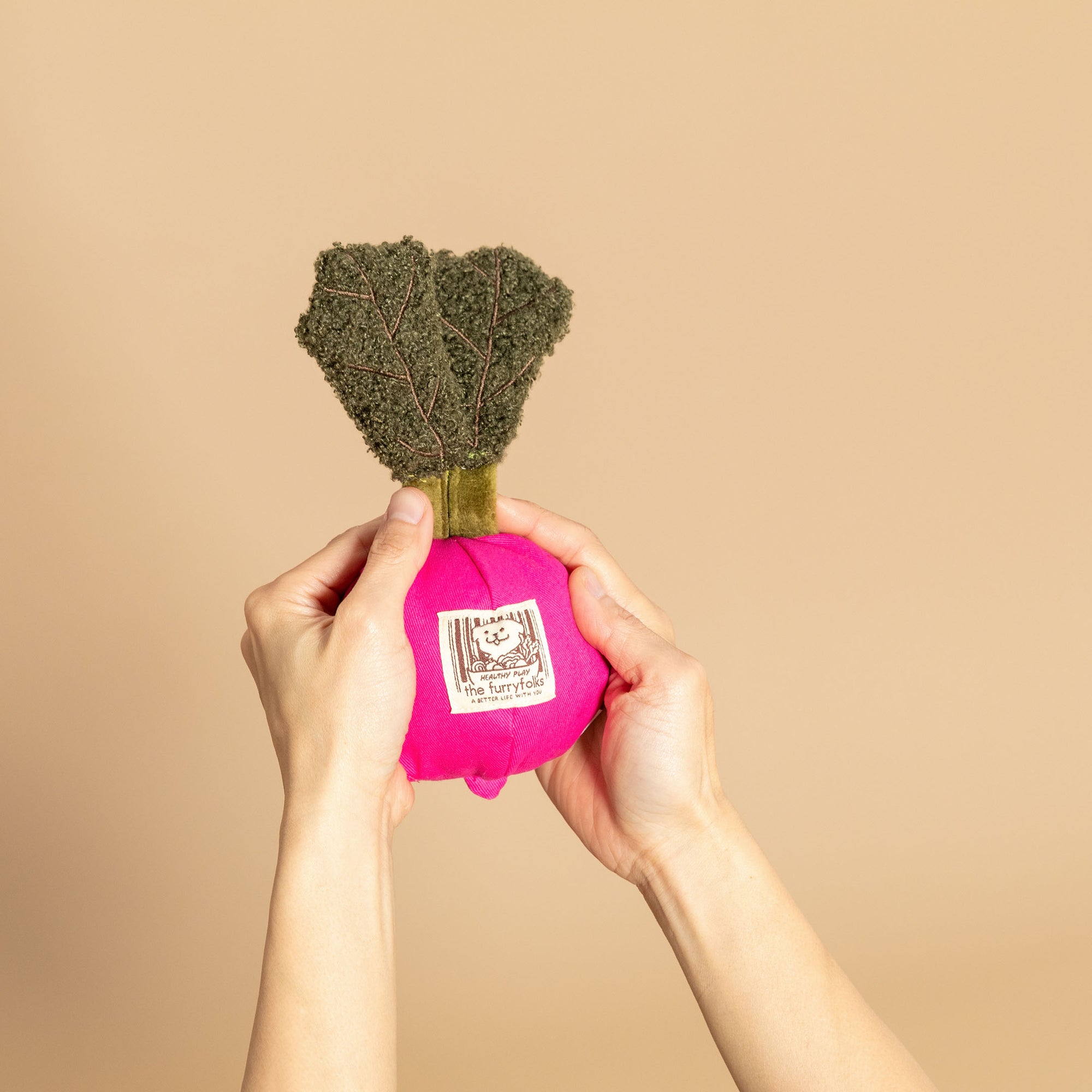 The image features two hands holding up a plush pink radish-shaped toy with a textured green top resembling leaves. The toy includes a label with "the furryfolks" branding, indicating it is part of a line of pet products. The beige background complements the toy's bright color, making it stand out as an attractive and playful item for dogs. The toy's design suggests it is intended for pets to enjoy both visually and tactilely, likely for activities like fetch or scent work.