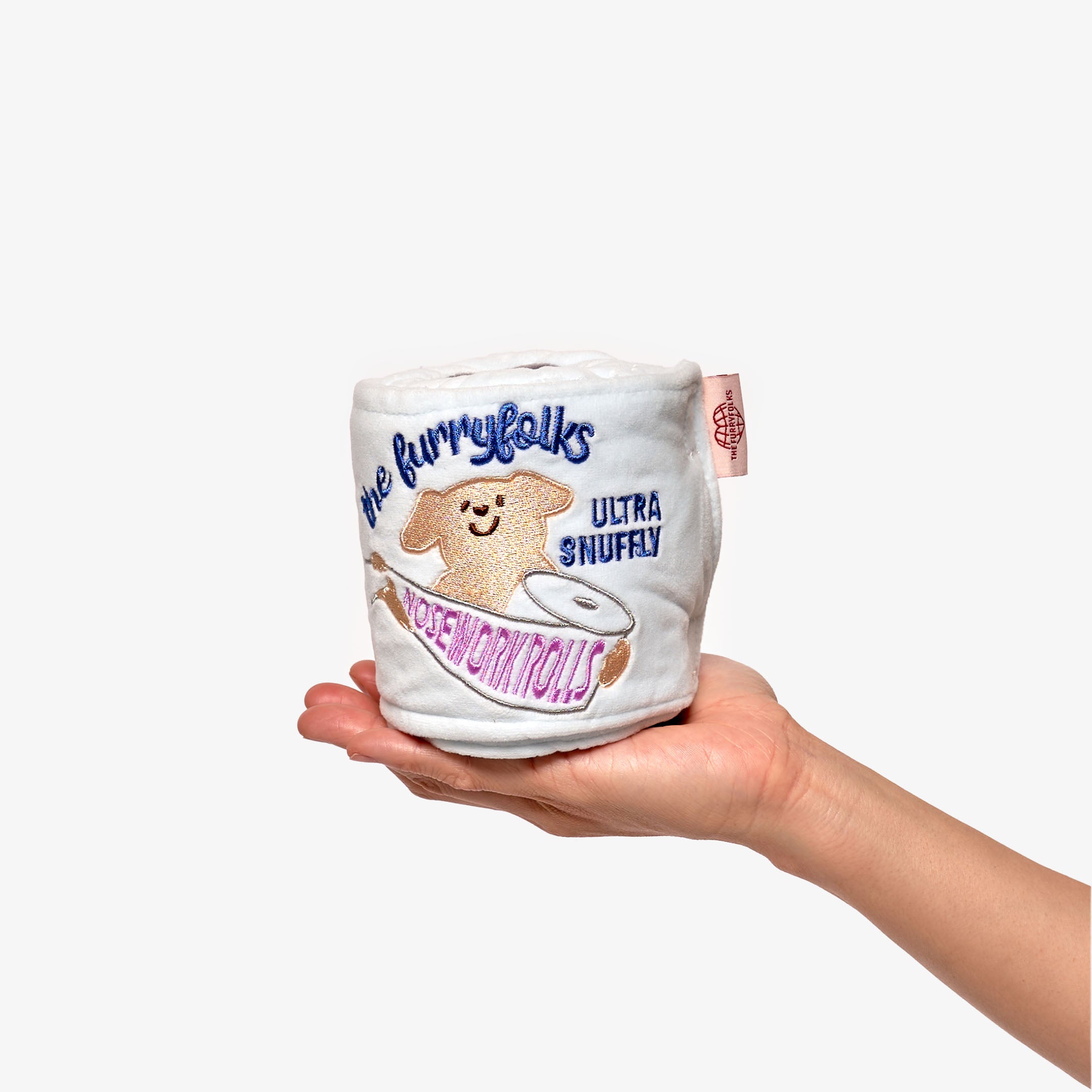The image shows a hand holding a plush dog toy. The toy is white with embroidered details including a smiling dog's face, the text "The Furryfolks", and the words "ULTRA SNUFFLY" in a decorative font. There's also a depiction of a toilet paper roll, adding a humorous touch. The toy has a pink tag on the side with what appears to be the brand's logo. The background is neutral, putting the focus on the toy itself. This appears to be a product designed for dogs to enjoy snuggling and playing with.