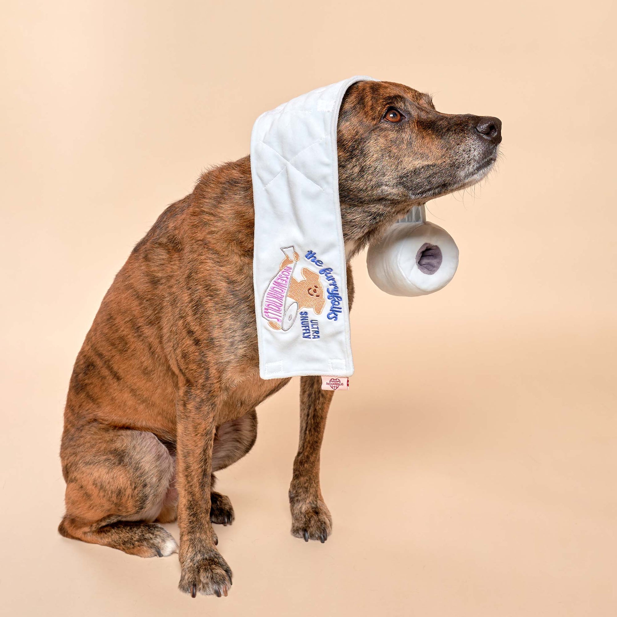 The image shows a brindle dog with a white snuffle mat draped over its neck, featuring "The Furryfolks" branding. The dog is looking to the side, giving the impression of being mid-movement or possibly distracted by something off-camera. The toy's rolled-up section suggests its use for hiding treats for the dog to find. The beige background emphasizes the dog and the toy, highlighting their interaction.