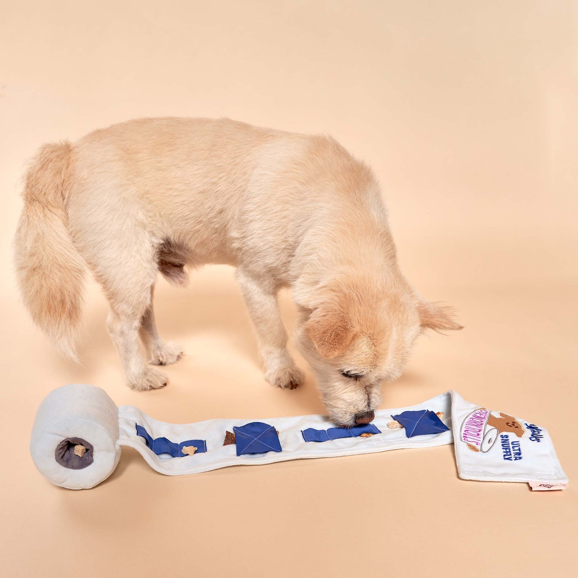 The image shows a light-colored dog engaging with a snuffle mat toy, which is laid out on the floor. The mat is white with blue pockets and has "The Furryfolks" branding on it, indicating it's designed for pets to use their sense of smell to find treats. The dog appears to be sniffing or retrieving a treat from one of the pockets, demonstrating the mat's intended use. The beige background complements the scene, highlighting the interaction between the dog and the toy.