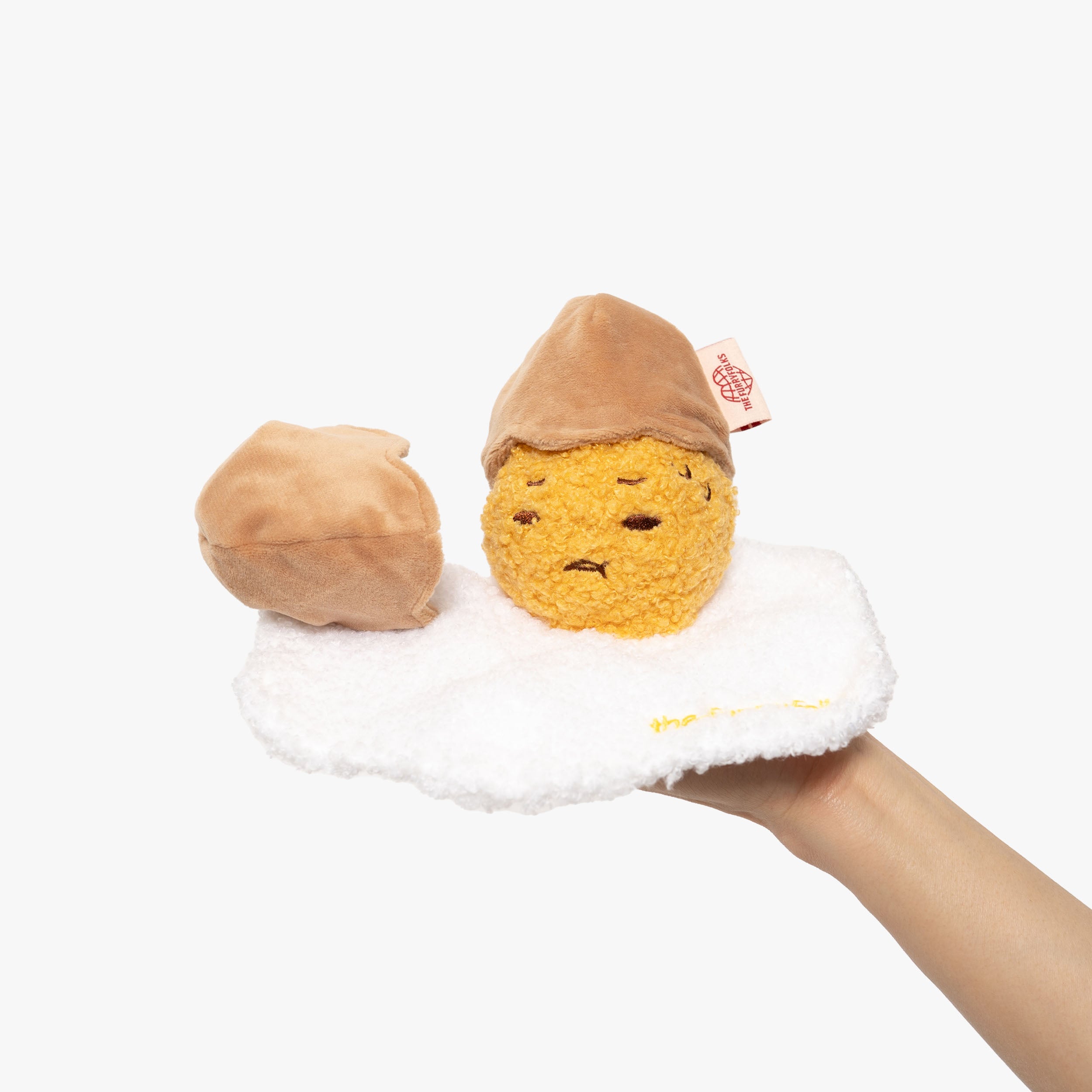 Adorable plush dog toy set, featuring a sunny-side-up egg with a textured yellow center and fluffy white border. Accompanied by a soft brown 'eggshell' hat, the set stimulates pets’ playtime with its interactive design. The toy encourages sniffing and foraging skills, perfect for engaging your furry friend in a healthy and fun activity. Bright, eye-catching, and crafted with care, it's a charming addition to any dog's toy collection. Tagged with 'The Furryfolks' for brand recognition.