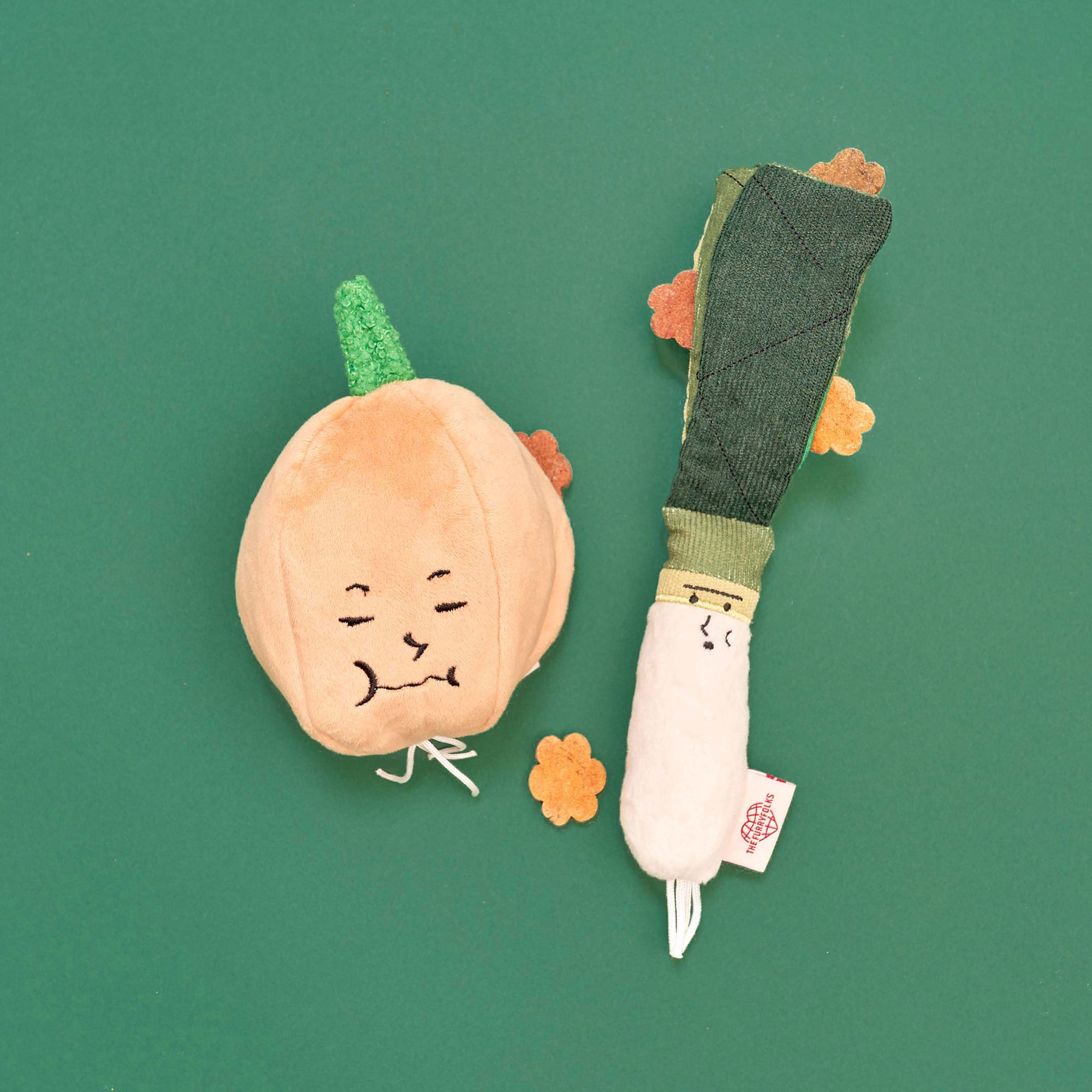 two plush dog toys, one shaped like a yellow onion with a happy face and green top, and another resembling a green onion with a content face and dark green leaves. Both are laid out on a green background with a few dog treats scattered around, indicating their function as interactive nosework toys that can be stuffed with treats to encourage a dog’s natural foraging behavior. The setup is simple yet visually engaging, highlighting the toys' features and potential for pet entertainment.