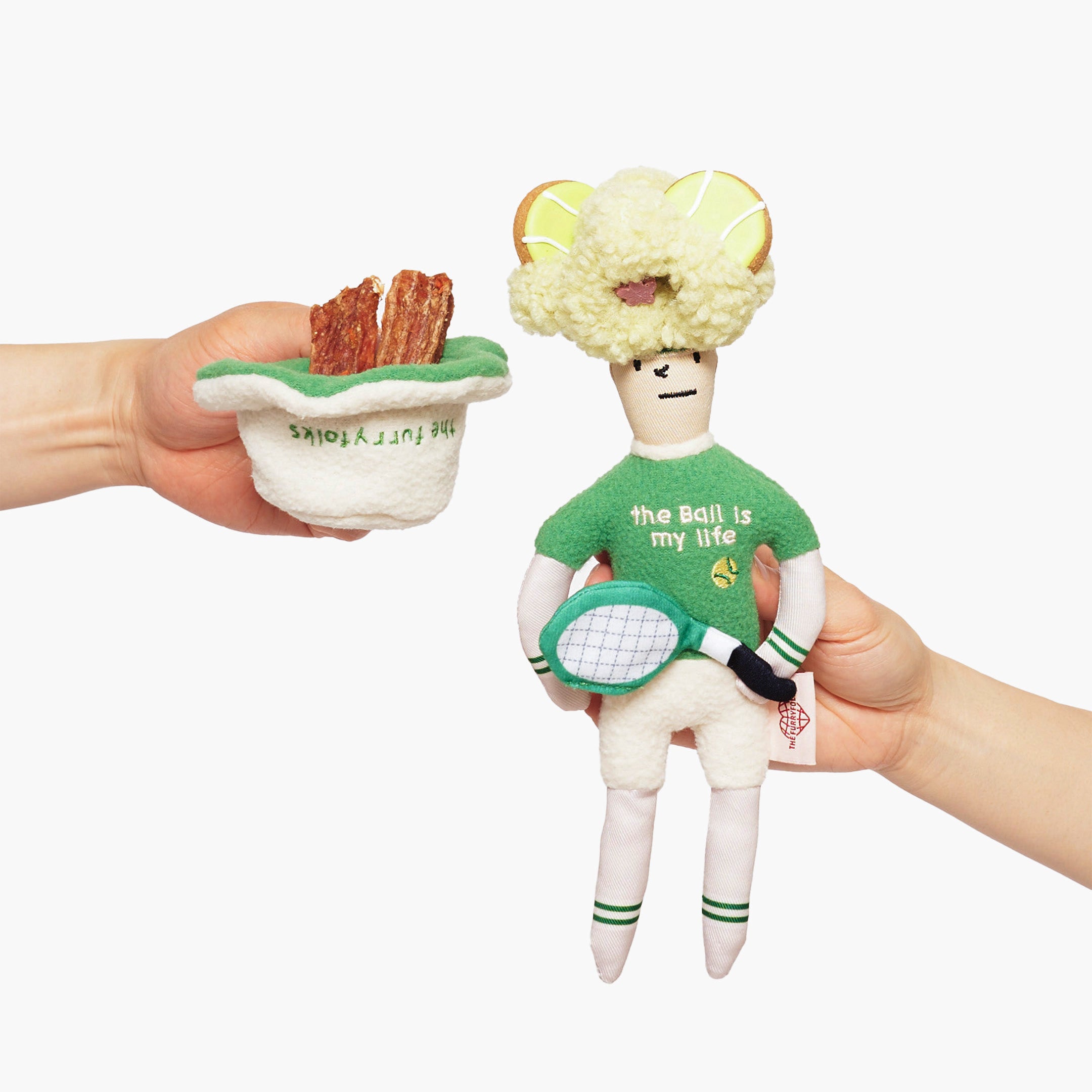 The image features two hands holding plush toys. On the left, a hand holds a small, plush cap filled with treats. On the right, another hand has the same plush toy as before, designed to resemble a human-like figure dressed as a tennis player, with a whimsical green hat, green sweater with the phrase "the ball is my life," and a white tennis racket. The figure's hat has added yellow "tennis ball" treats. The design is playful and imaginative.