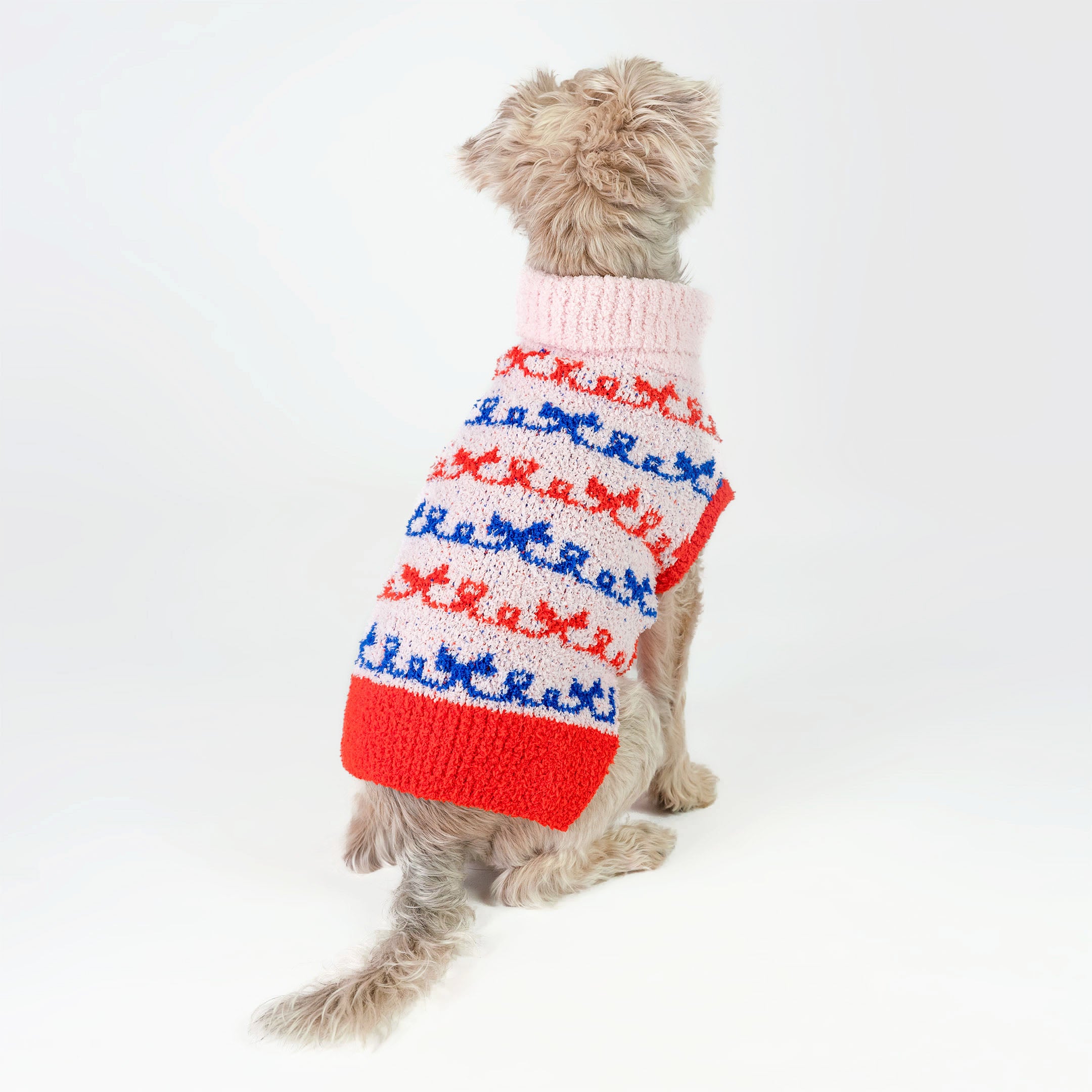 Small dog sitting with its back turned, wearing a pink sweater with red and blue bow patterns. The cozy knit design offers both warmth and style, showcasing a fun and colorful look perfect for any furry friend.