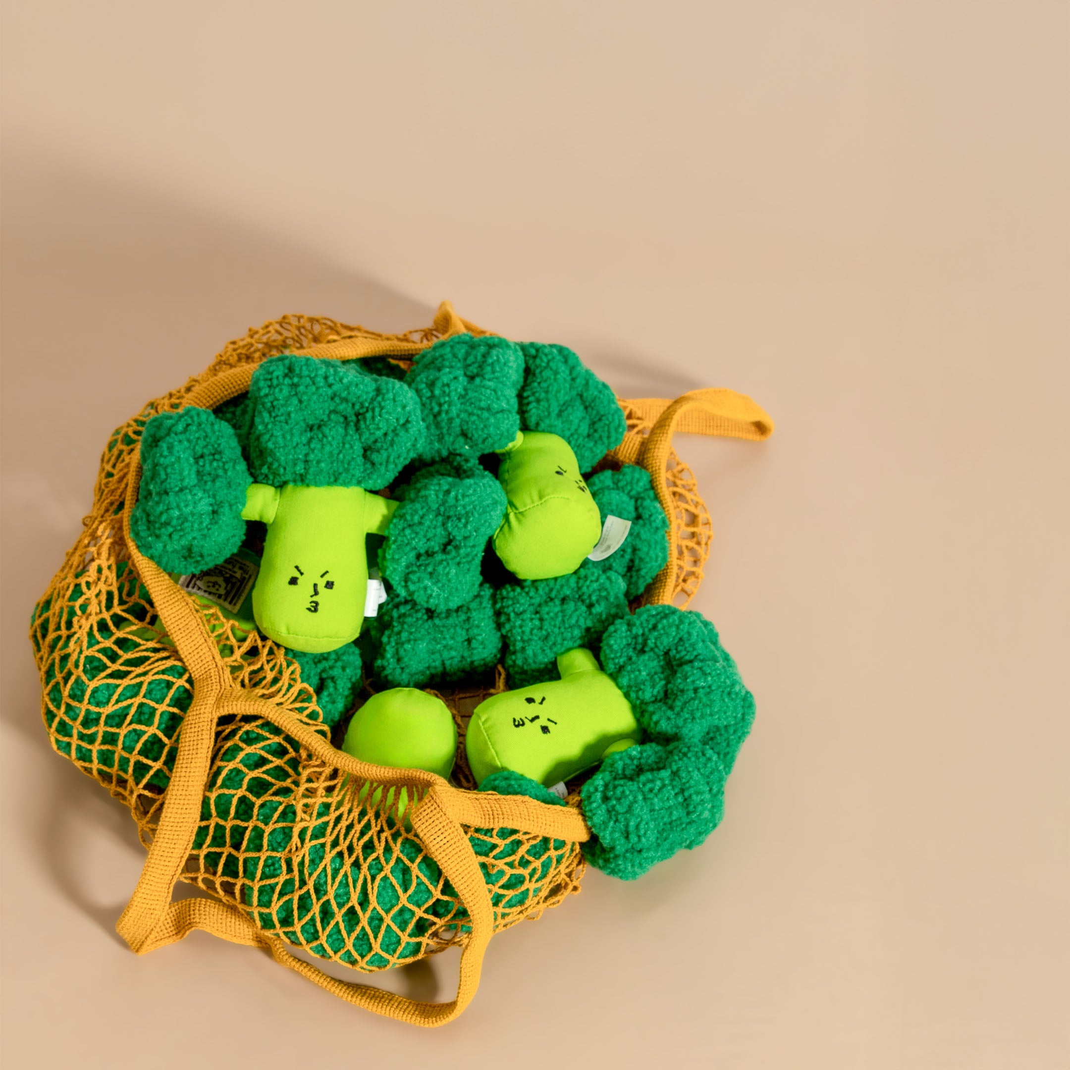 Plush broccoli toys in a yellow mesh bag on a beige background