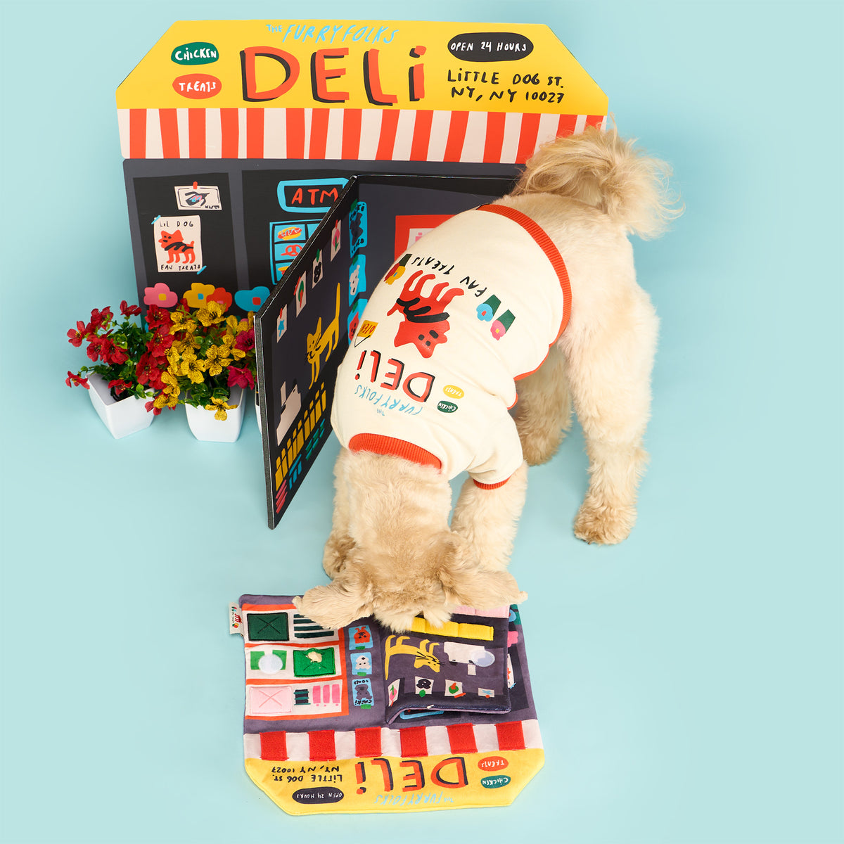 Cream-colored dog wearing a deli-print shirt sniffs the open Deli Nosework mat on a light blue floor beside a matching storefront backdrop.
