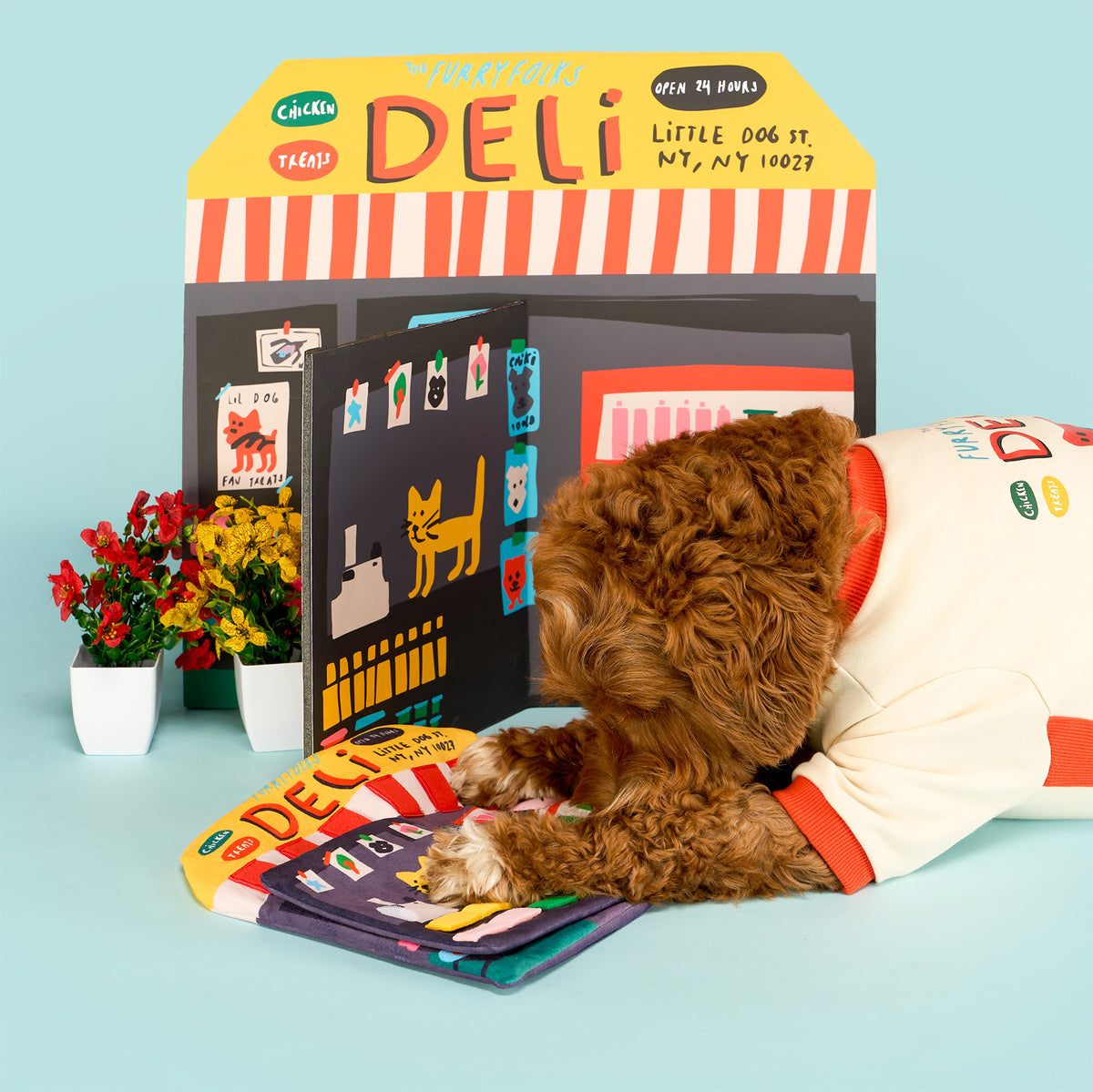 Brown curly-haired dog in a deli-print shirt lies with its paws on the open Deli Nosework mat, sniffing its fabric pockets.
