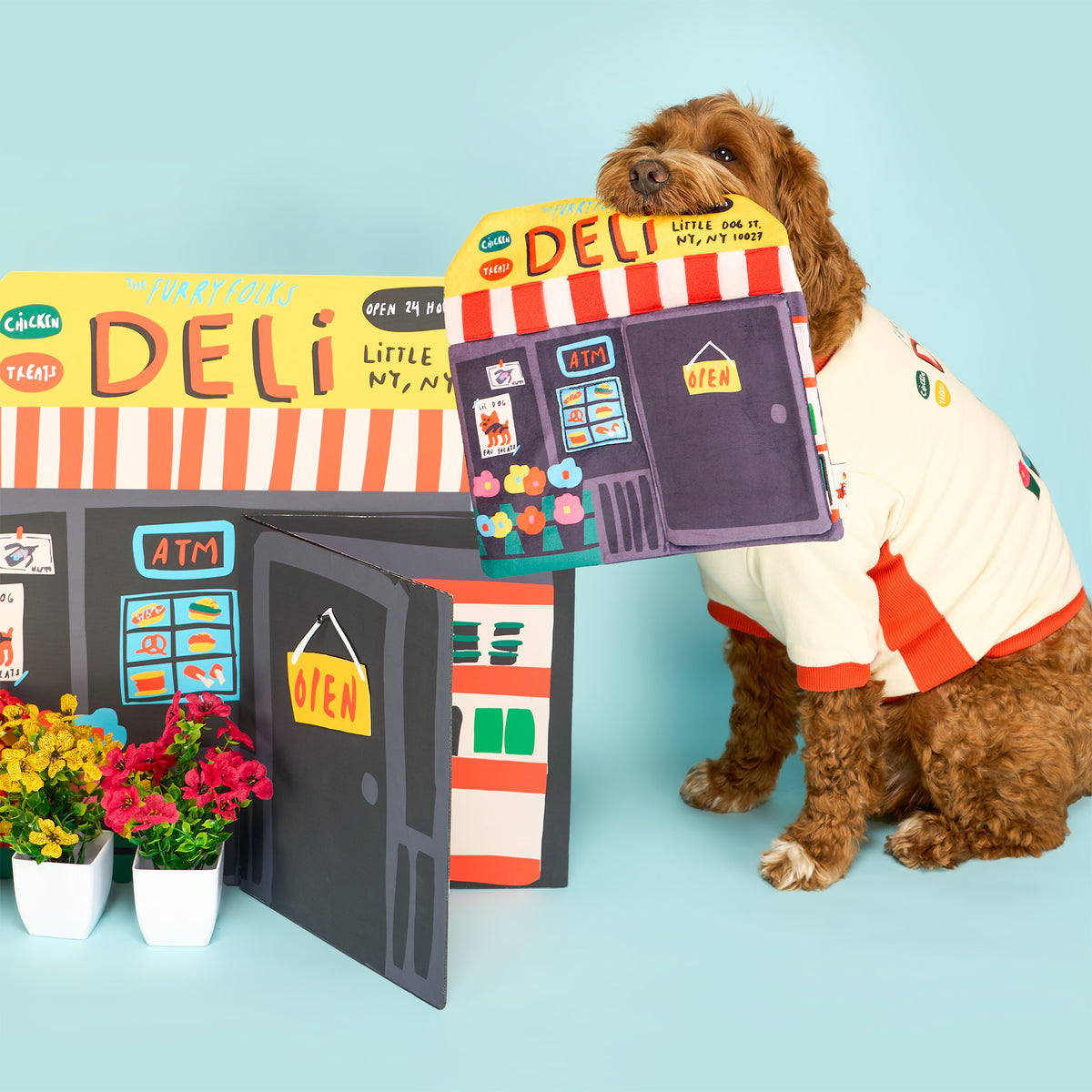 Brown dog in a cream deli-print shirt holds the Deli Nosework mat in its mouth beside a matching storefront display and colorful flowers.