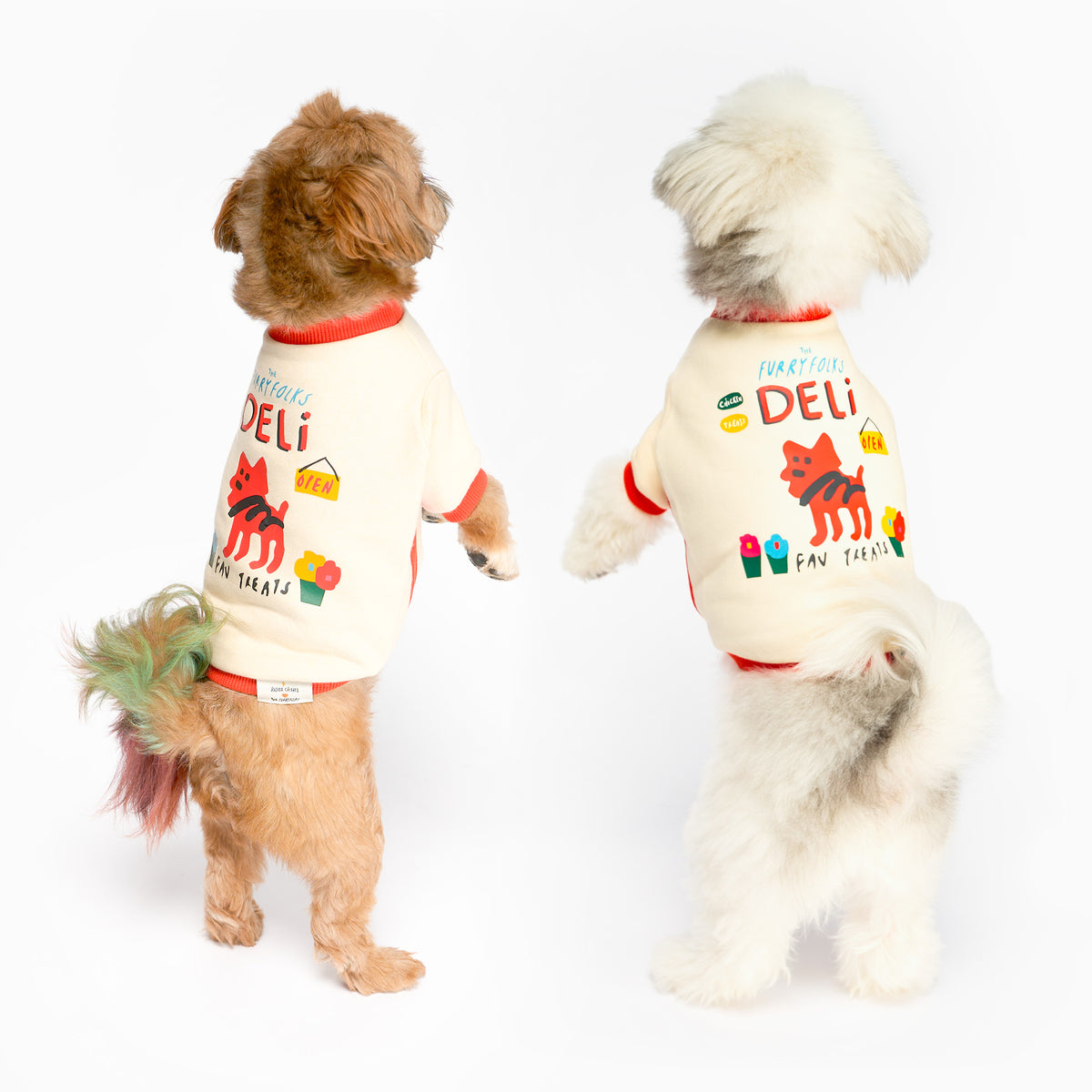 Two small dogs, one brown and one white, stand on their hind legs with their backs to the camera, wearing matching cream shirts with red trim, colorful dog and flower graphics, and “the furryfolks Deli” lettering.