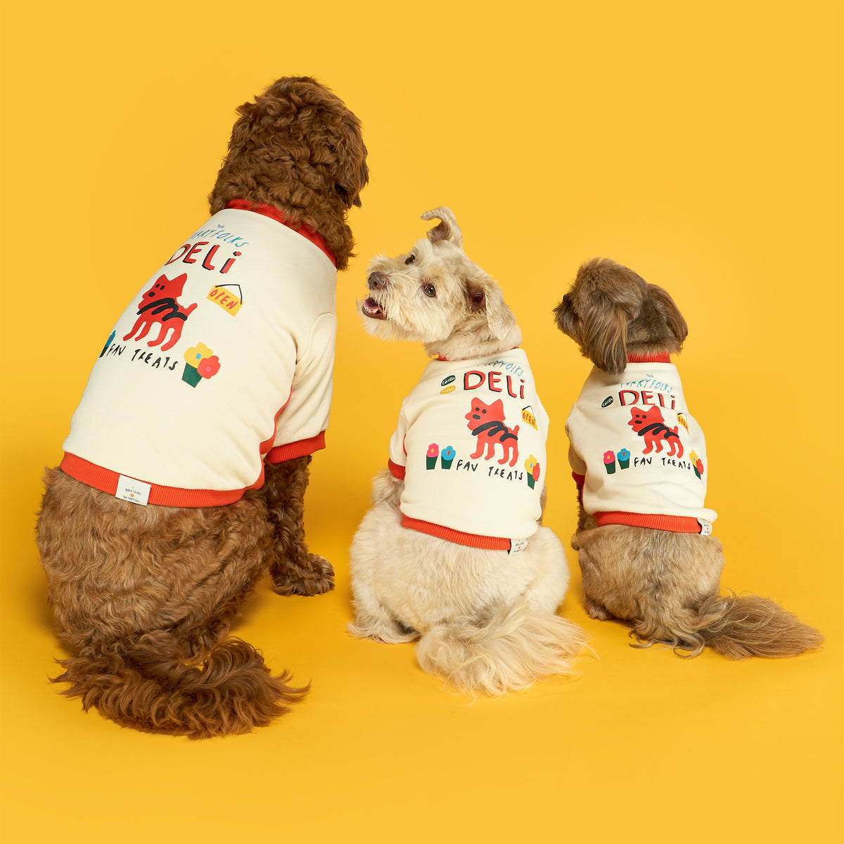 Three dogs of different sizes sit with their backs to the camera against a yellow background, wearing matching cream “the furryfolks Deli” shirts with orange trim and colorful graphics. The middle dog looks back over its shoulder.