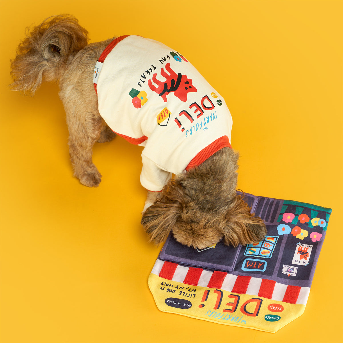 Overhead view of a small brown dog wearing a cream “the furryfolks deli” shirt with orange trim, sniffing a colorful deli storefront-shaped fabric toy on a bright yellow floor.