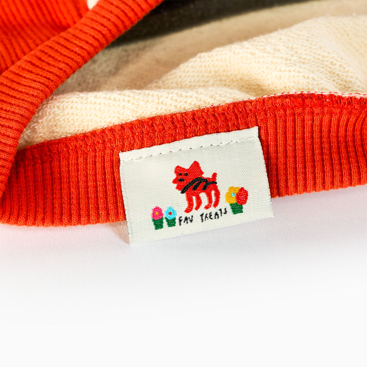 Close-up of orange ribbed trim and cream looped fabric on a dog shirt, with a woven label featuring a red cartoon dog, colorful potted flowers, and “fav treats” lettering.