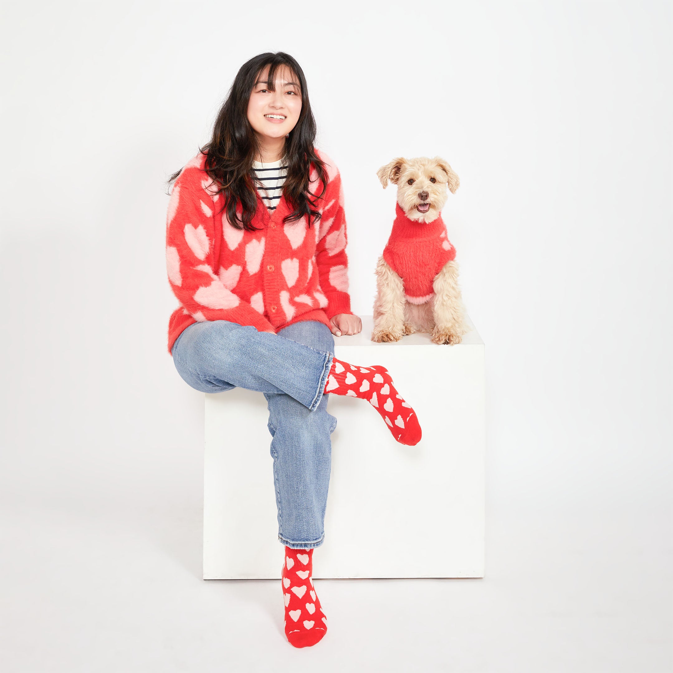 A woman sitting on a white cube, wearing a red cardigan with pink hearts, paired with red socks with white hearts. A small beige dog sits next to her, wearing a matching red sweater with pink heart patterns.