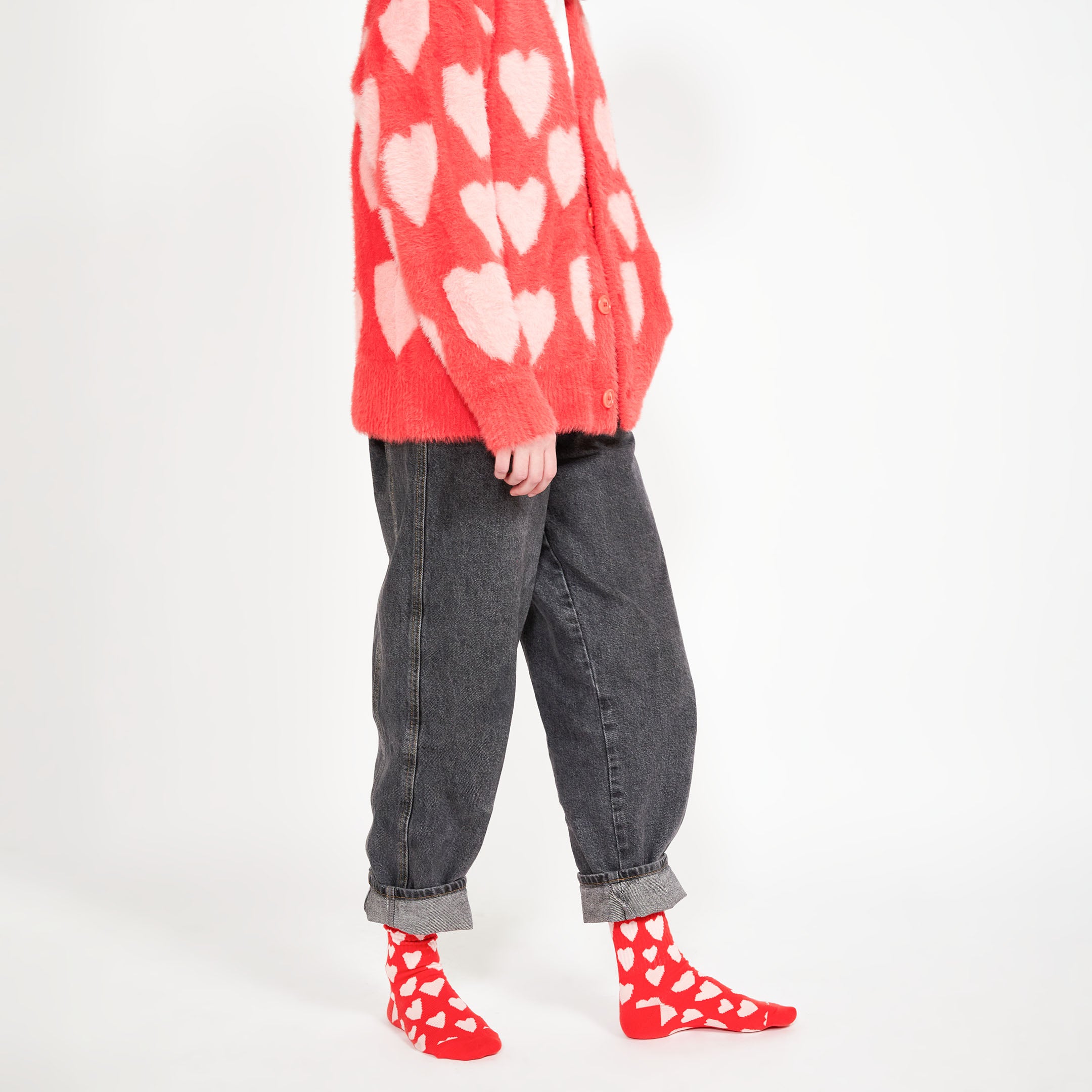  A woman standing with rolled-up jeans, red socks with white hearts, and a red cardigan with pink hearts, showing off a playful and cozy look.