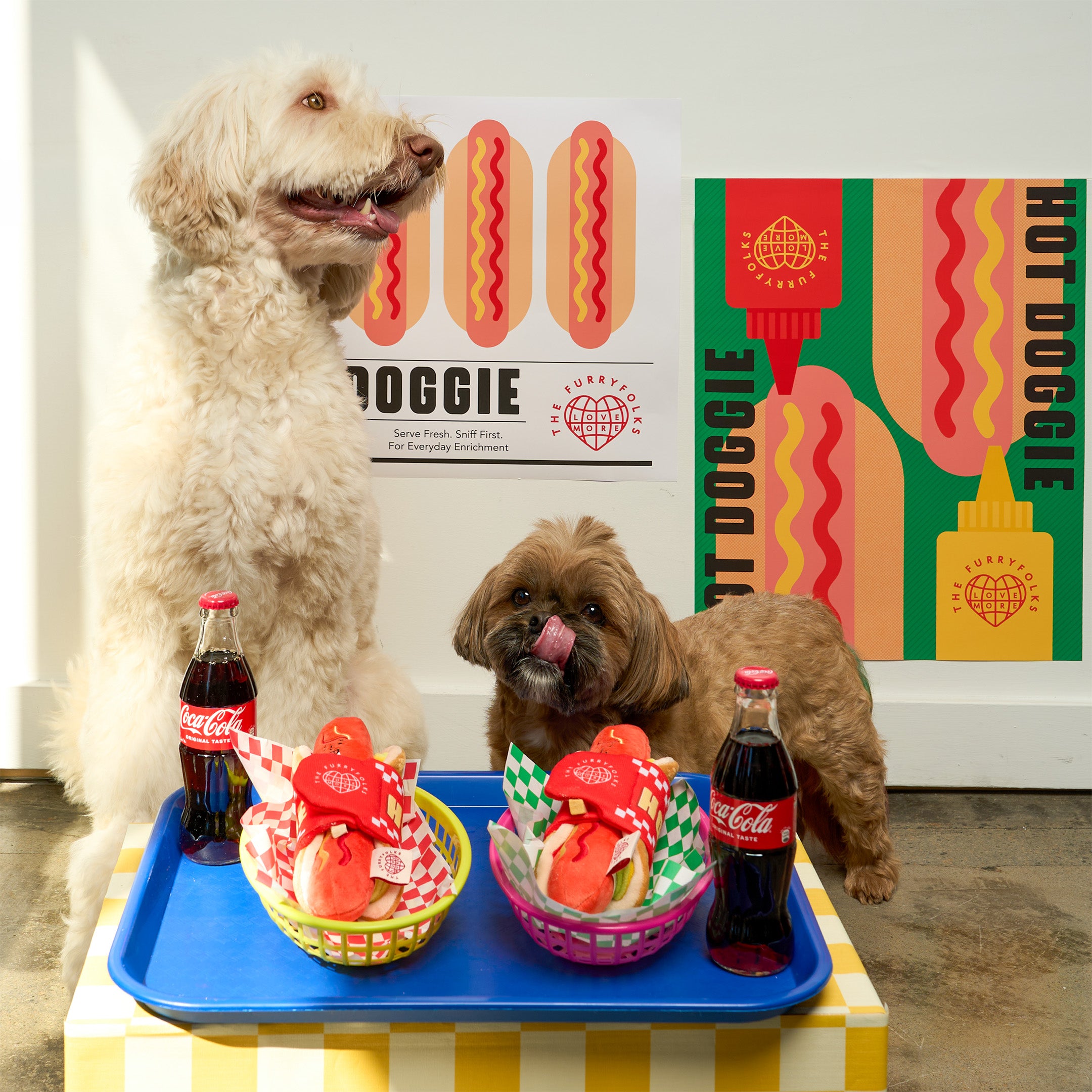 Two dogs with hot dog treats and Coca-Cola bottles on a tray, with a colorful 'Hot Doggie' poster in the background.
