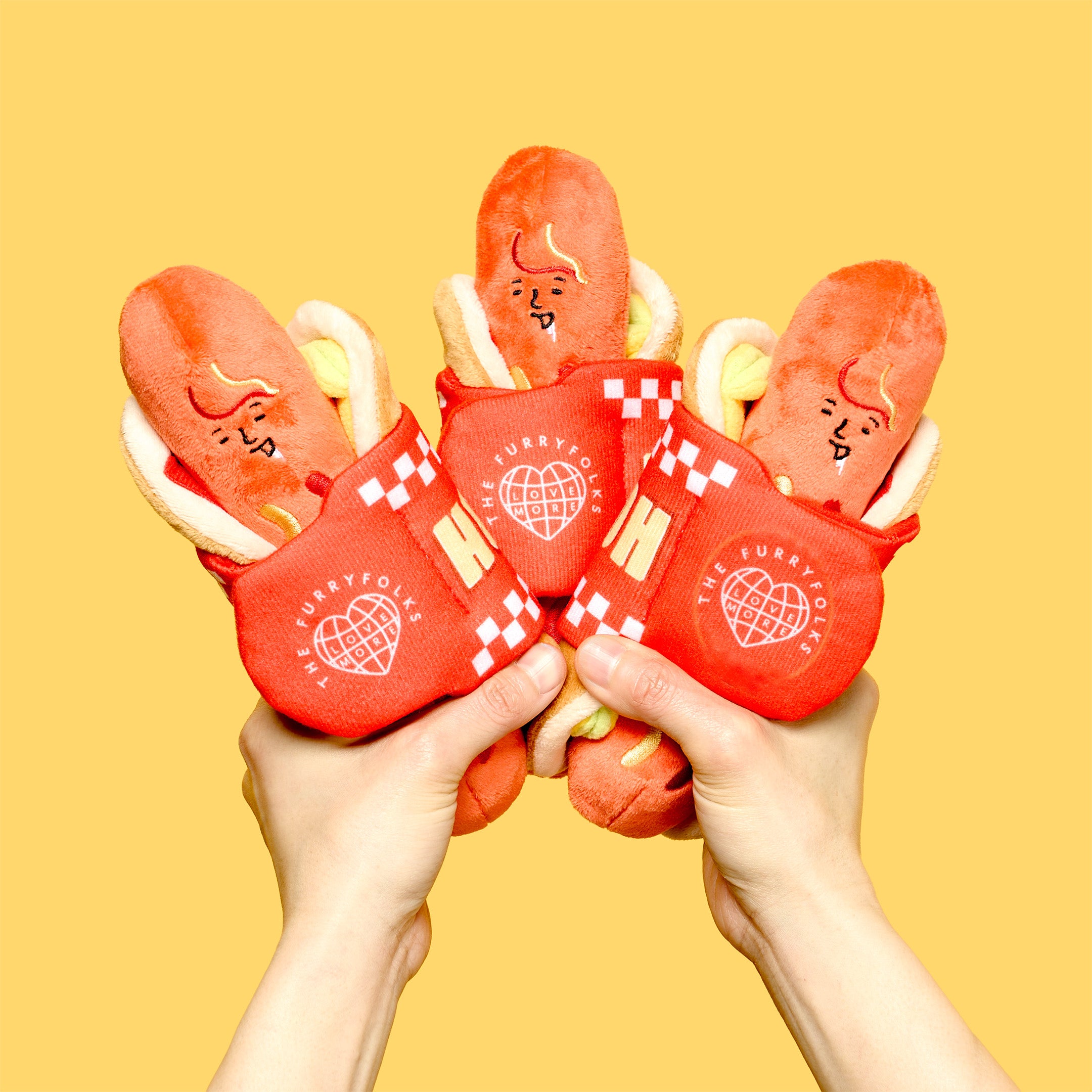 Red plush hot dogs with checkered buns held by hands against a yellow background