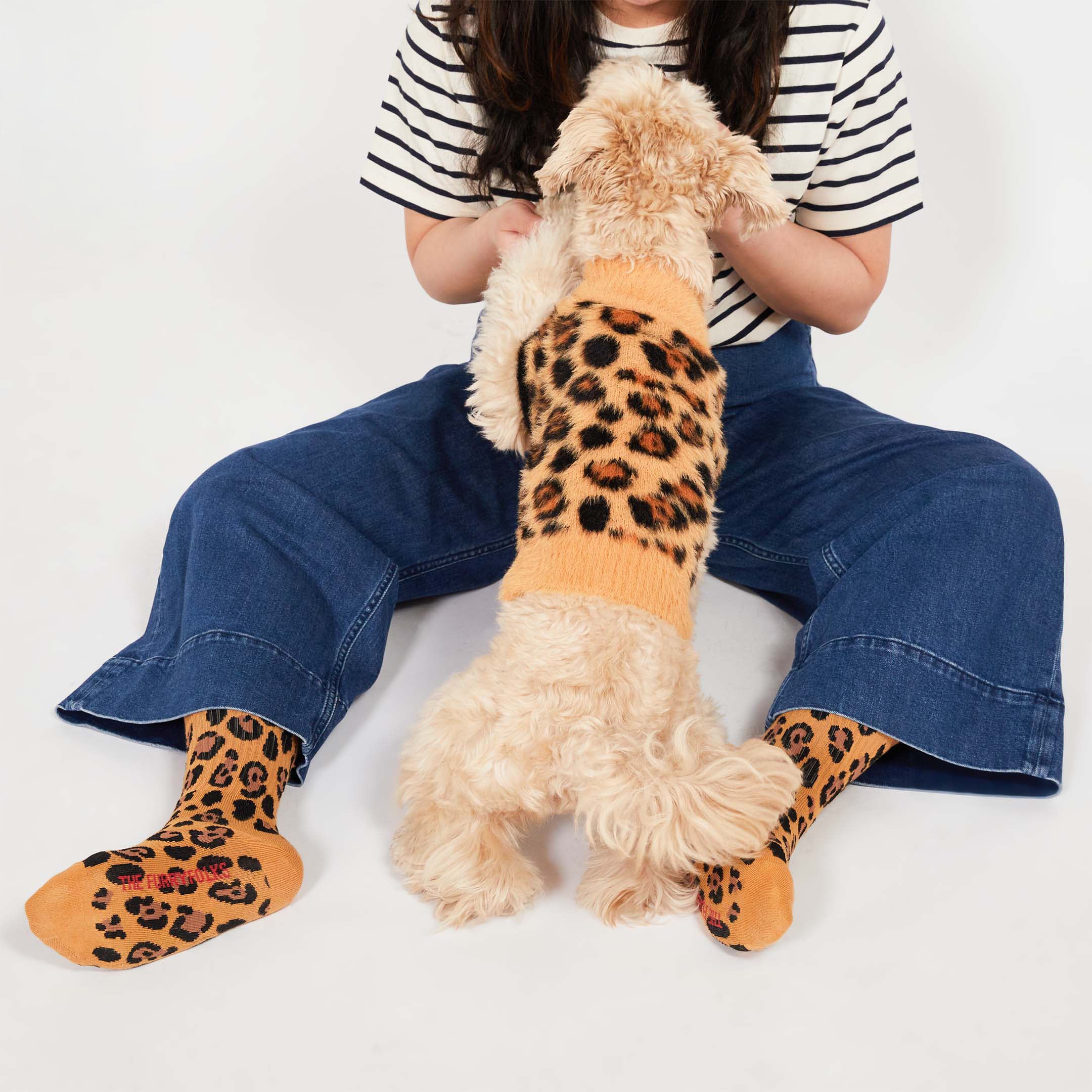 A light-colored dog wearing a leopard-print sweater stands on its hind legs, facing a person seated on the ground. The person is wearing a striped shirt, blue flared jeans, and leopard-print socks.