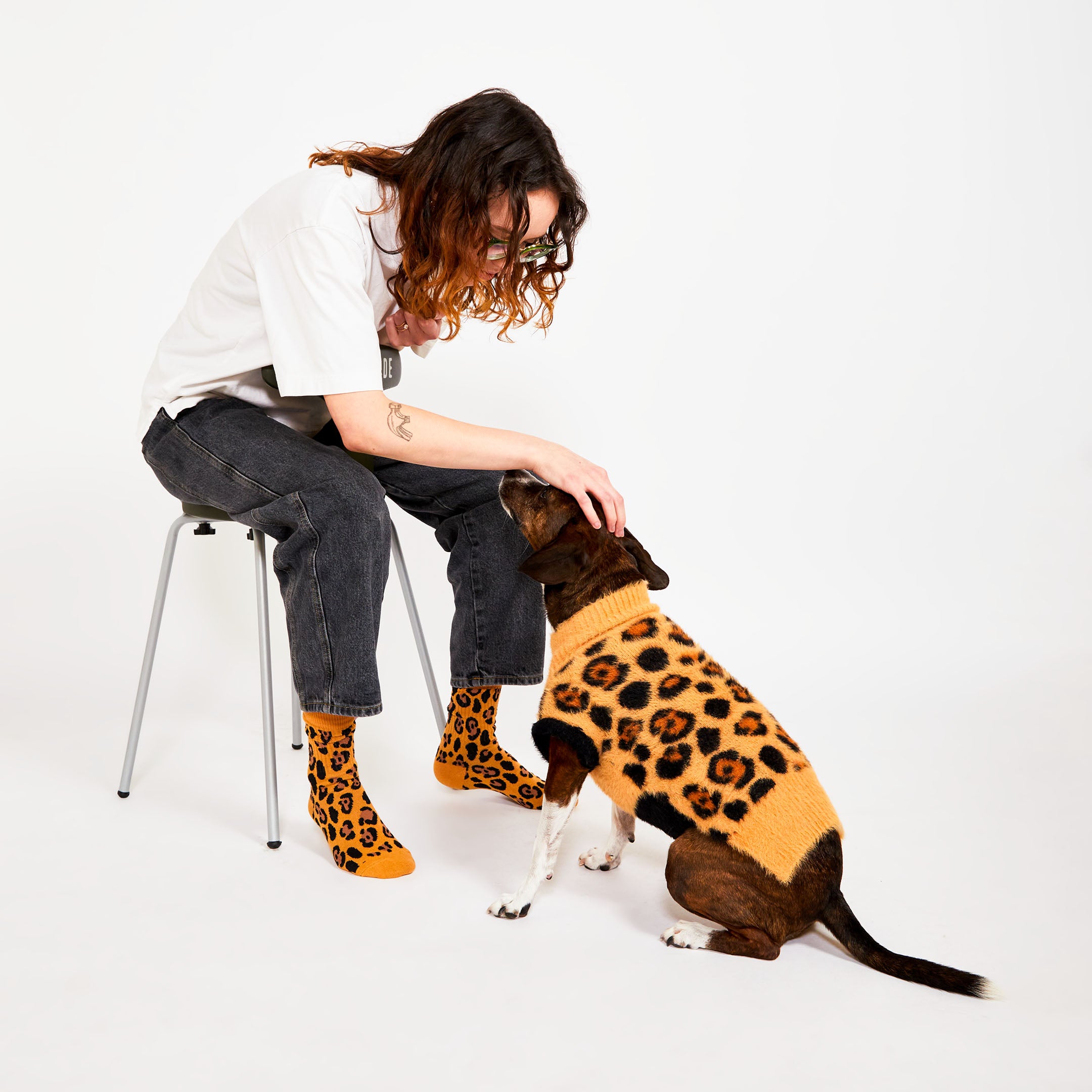 A  person with shoulder-length hair, sitting on a stool, leans down to pet a dog wearing a leopard-print sweater. The person is dressed casually in black jeans and leopard-print socks, matching the dog's outfit.