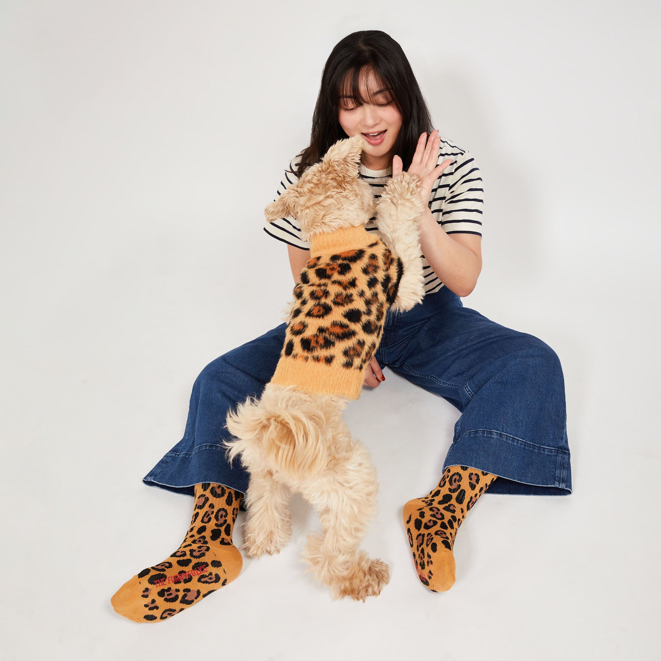 A light-colored dog wearing a leopard-print sweater stands on its hind legs, high-fiving a woman sitting on the ground. The woman is wearing a striped shirt, flared blue jeans, and matching leopard-print socks.