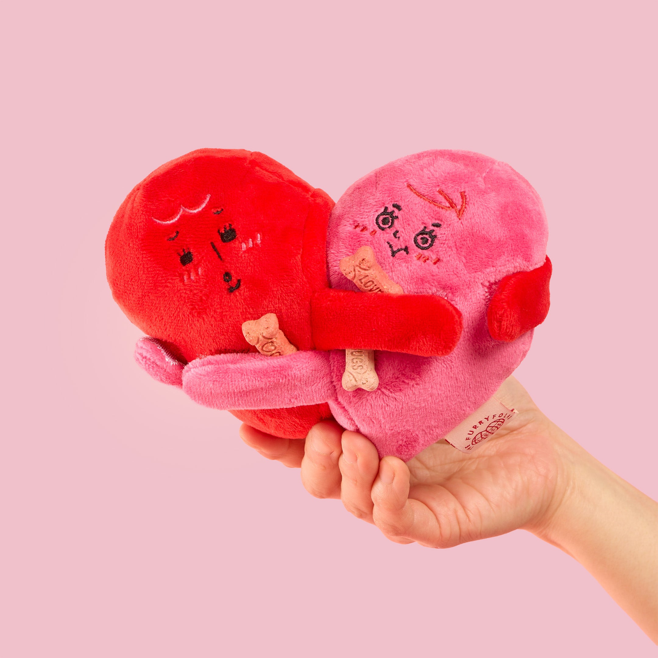 Heart-shaped plush toys held in a hand against a pink background