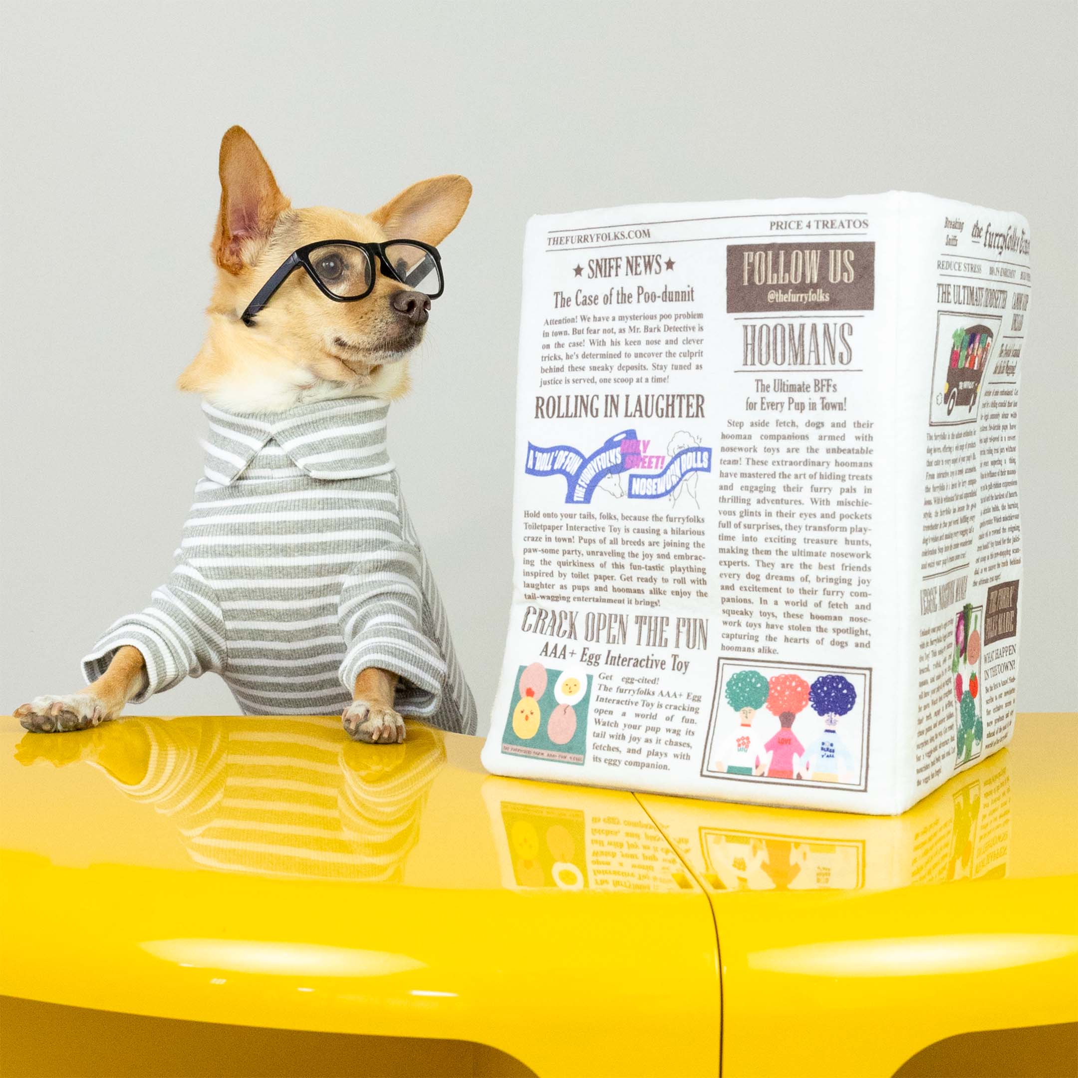 A Chihuahua in glasses and a striped shirt stands on a yellow table, leaning on a folded newspaper-themed toy. The dog poses as if reading, further adding to the playful and quirky atmosphere.