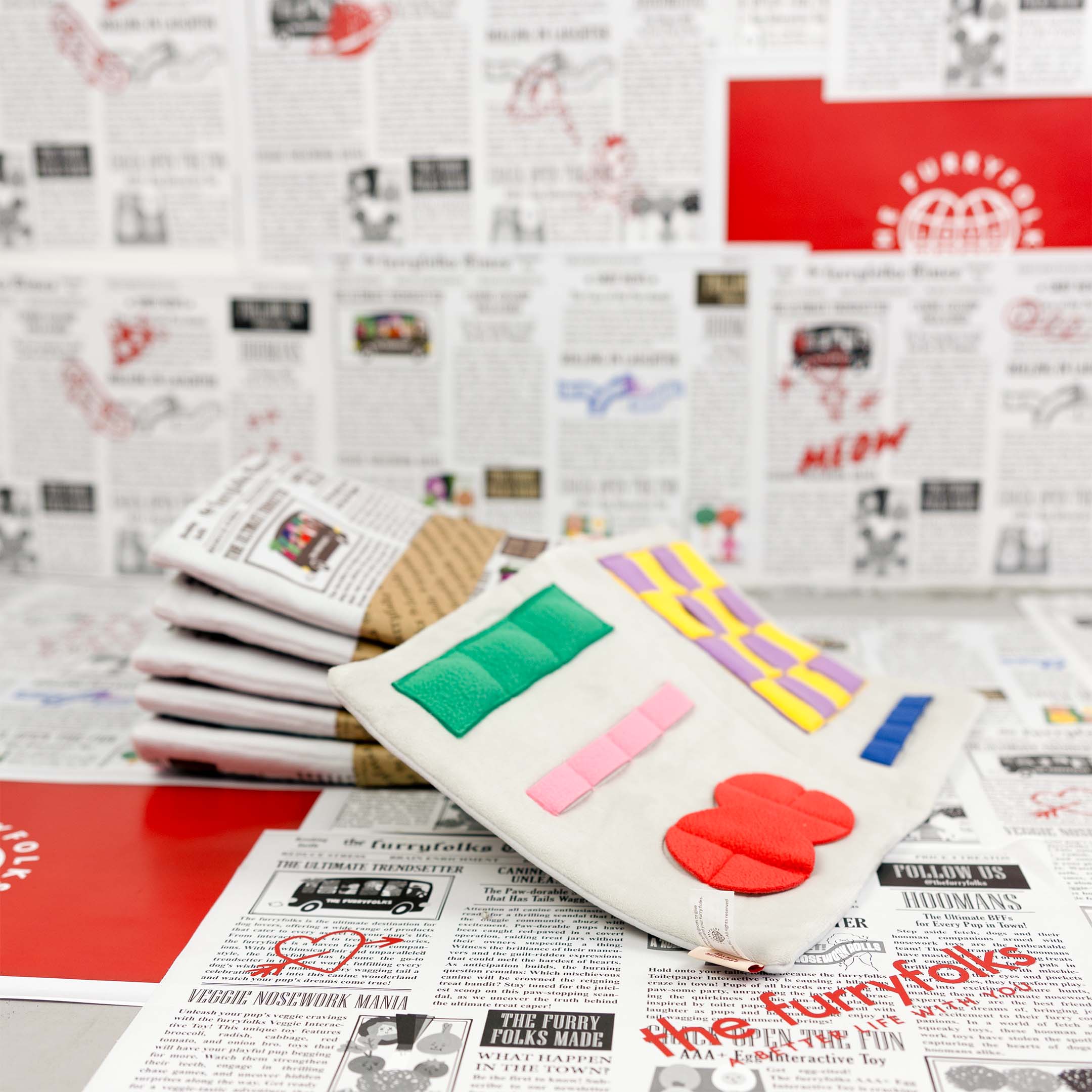 A stack of newspaper-themed dog toys is shown in the foreground, with an open nosework toy on top displaying colorful compartments. The background is decorated with playful newspaper designs.