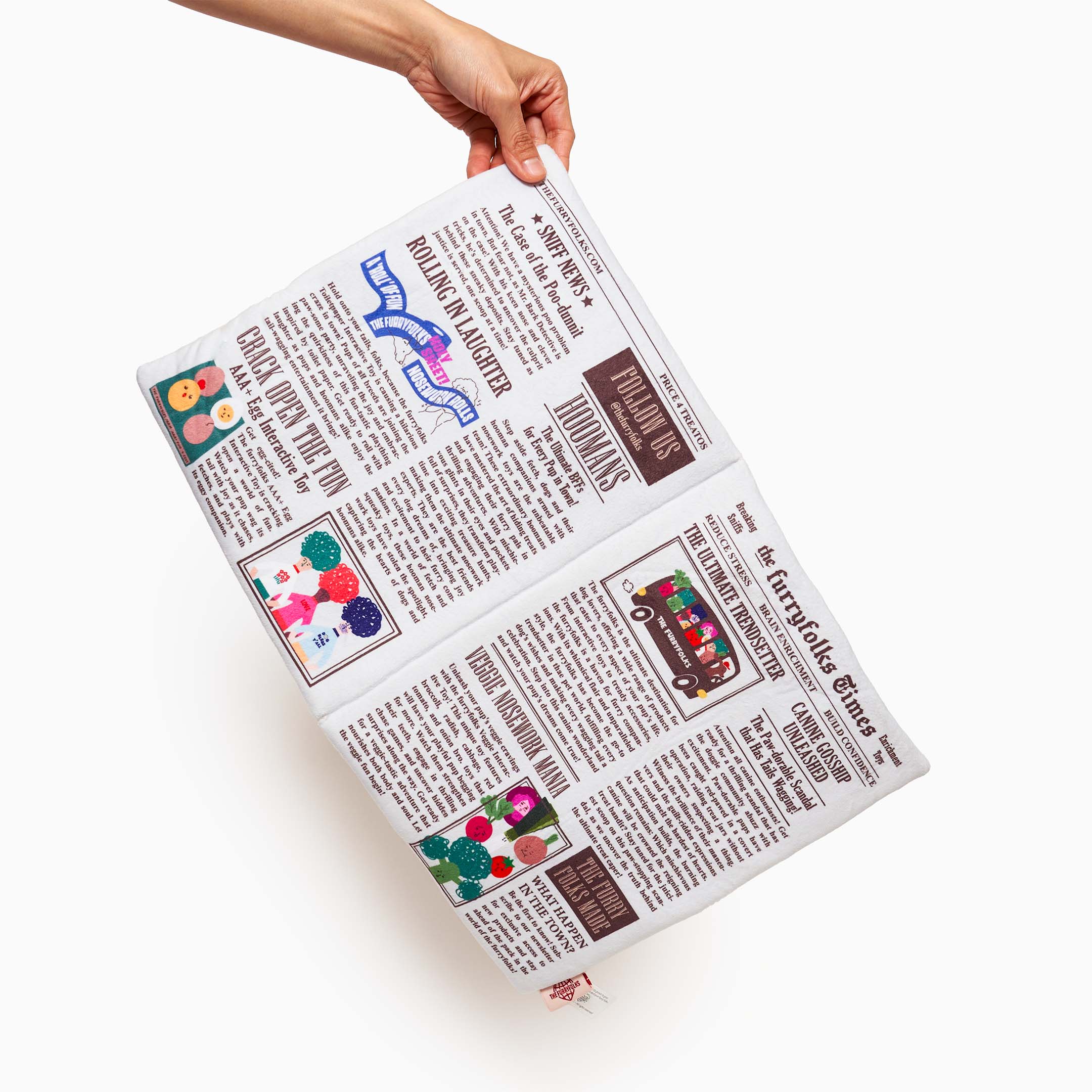 A hand holding a pet nosework toy designed to resemble a newspaper. The fabric features printed articles, playful illustrations, and humorous headlines like 'Rolling in Laughter' and 'the furryfolks Times.' Each section mimics the look of a newspaper page, providing an engaging and interactive experience for pets as they sniff and search for hidden treats within the folds and pockets.