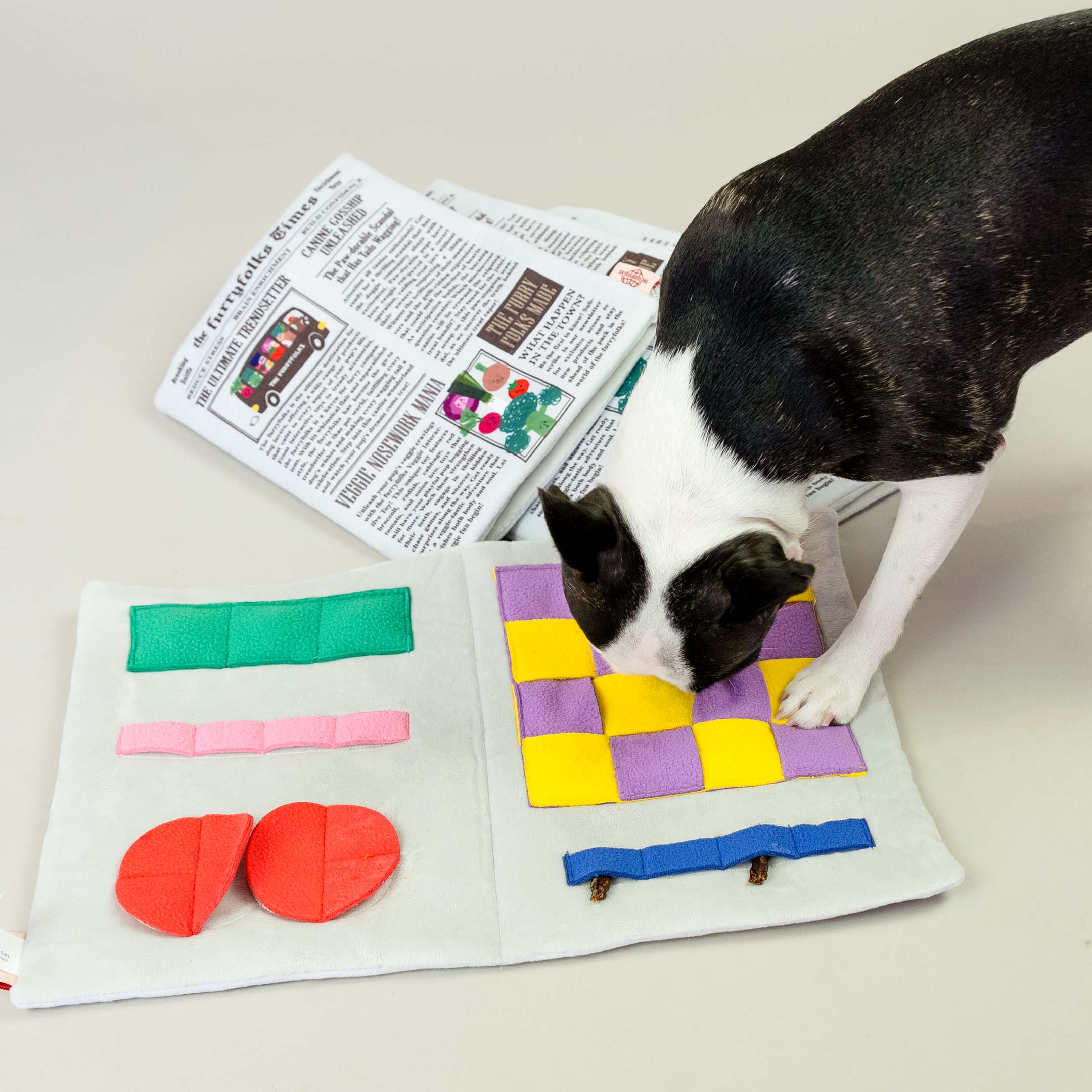 This second image shows the same dog interacting with the nosework toy, which features a more detailed view of the toy’s inner design. The open toy has various compartments, likely intended to hide treats or other small items for the dog to find. The colorful fabric sections, including the green, pink, red, yellow, and purple squares, are likely designed to engage the dog's sense of smell and curiosity.