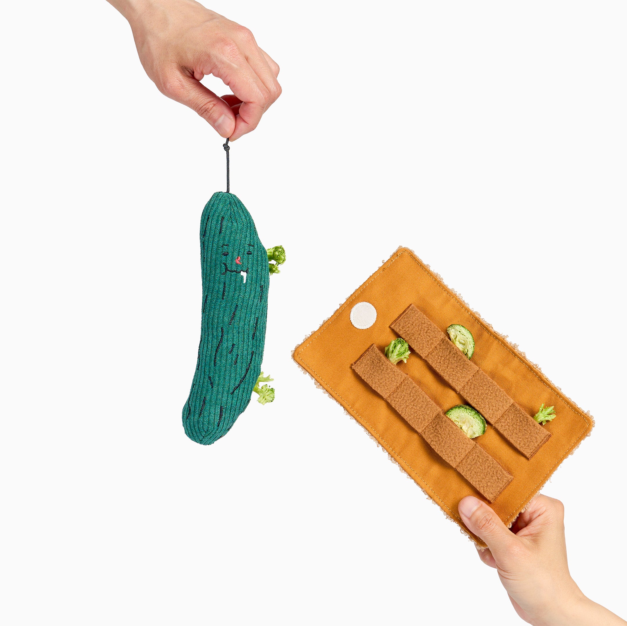 A green cucumber-shaped plush pet toy with a face is being held by a hand, while another hand holds an orange fabric training mat with brown straps and small cucumber pieces on a white background.