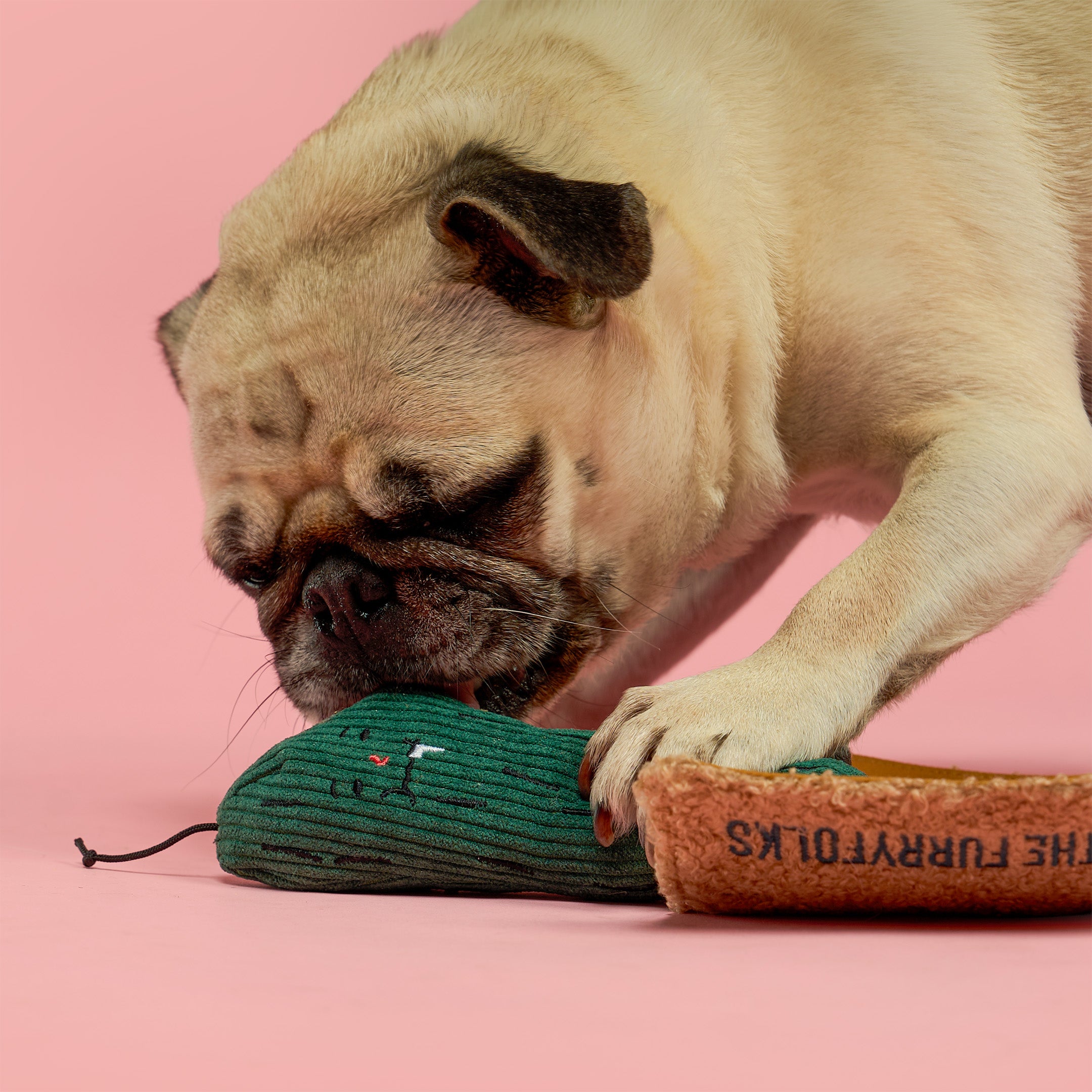 A pug dog playfully biting and chewing a green ribbed mouse-shaped toy next to a brown cardboard tube printed with 'THE FURRY TOYS KS' on a solid pink background.