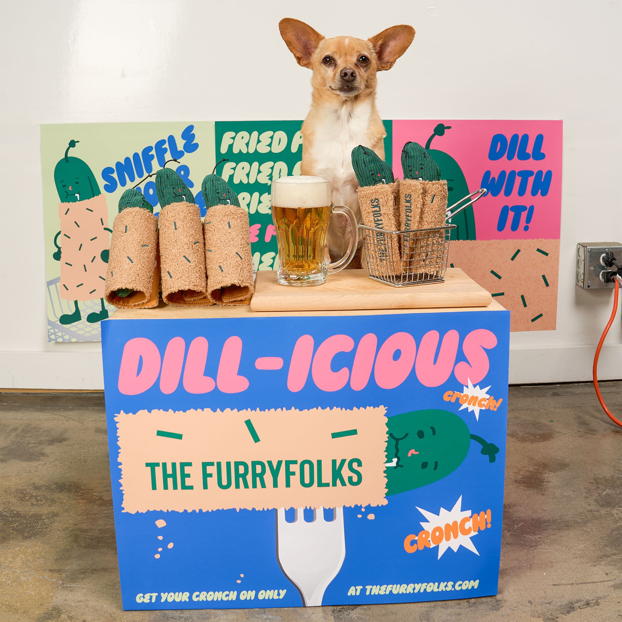 A small tan dog stands behind a branded blue display box labeled 'DILL-ICIOUS THE FURRYFOLKKS' with colorful dill-themed signs in the background. On top of the box are wrapped food items, a glass mug of beer, and a wire basket, all in a styled indoor setting.