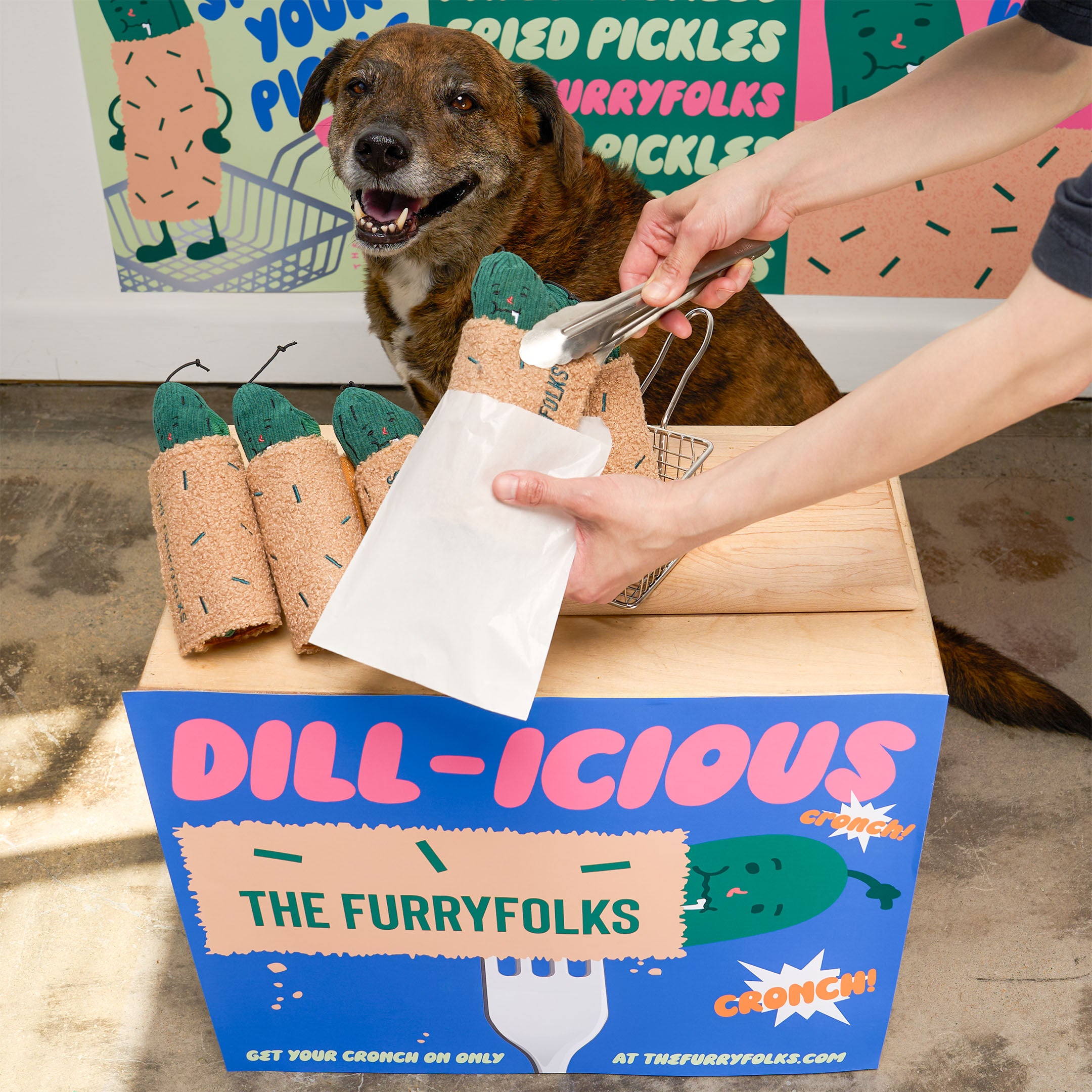 A smiling brindle dog sits behind a colorful “Dill-icious” market display as a vendor uses tongs to place a pickle-shaped plush dog toy into a paper bag.